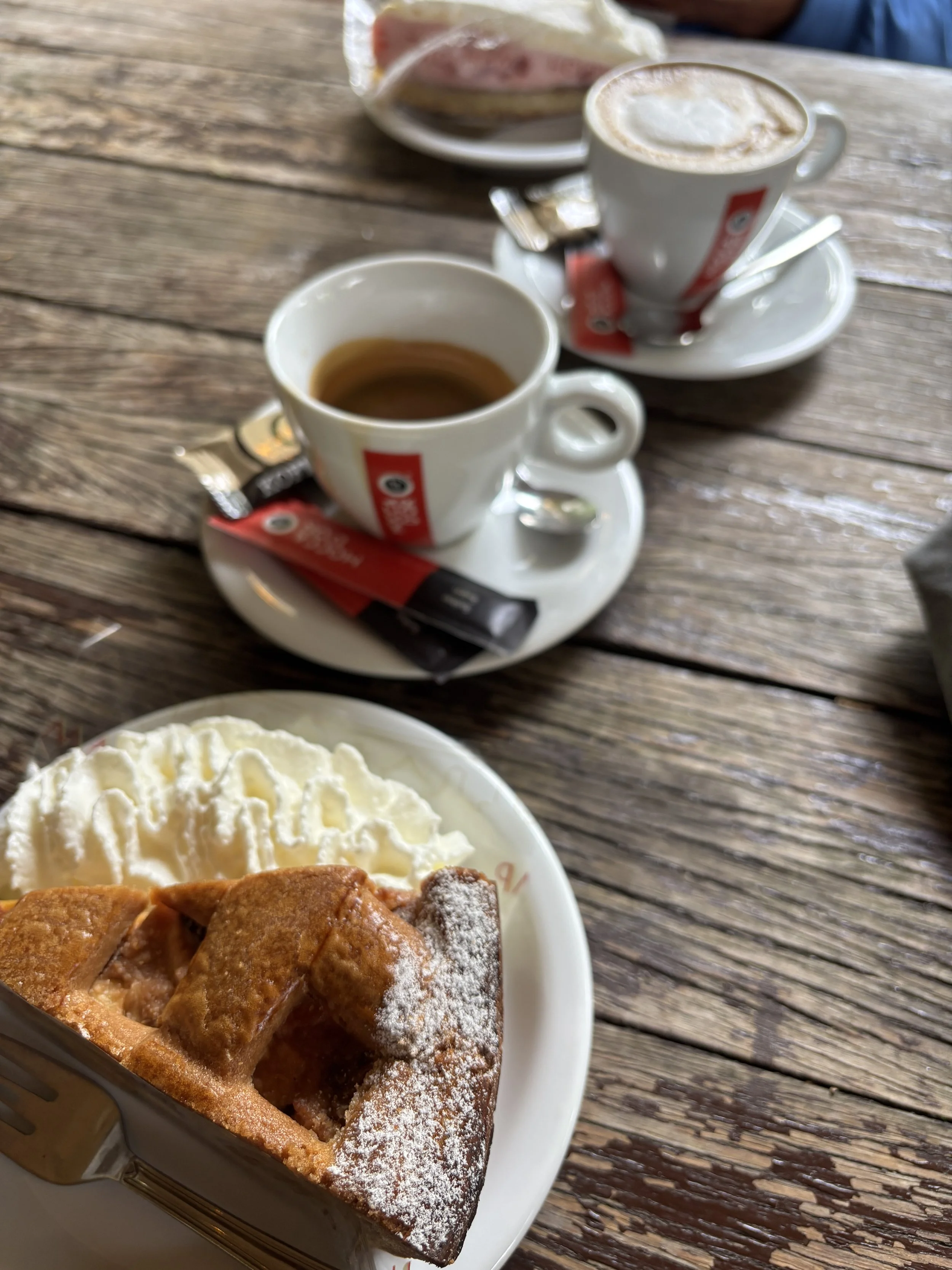 A wooden table with a plate of apple pie topped with powdered sugar and a serving of whipped cream, alongside cups of coffee or espresso.