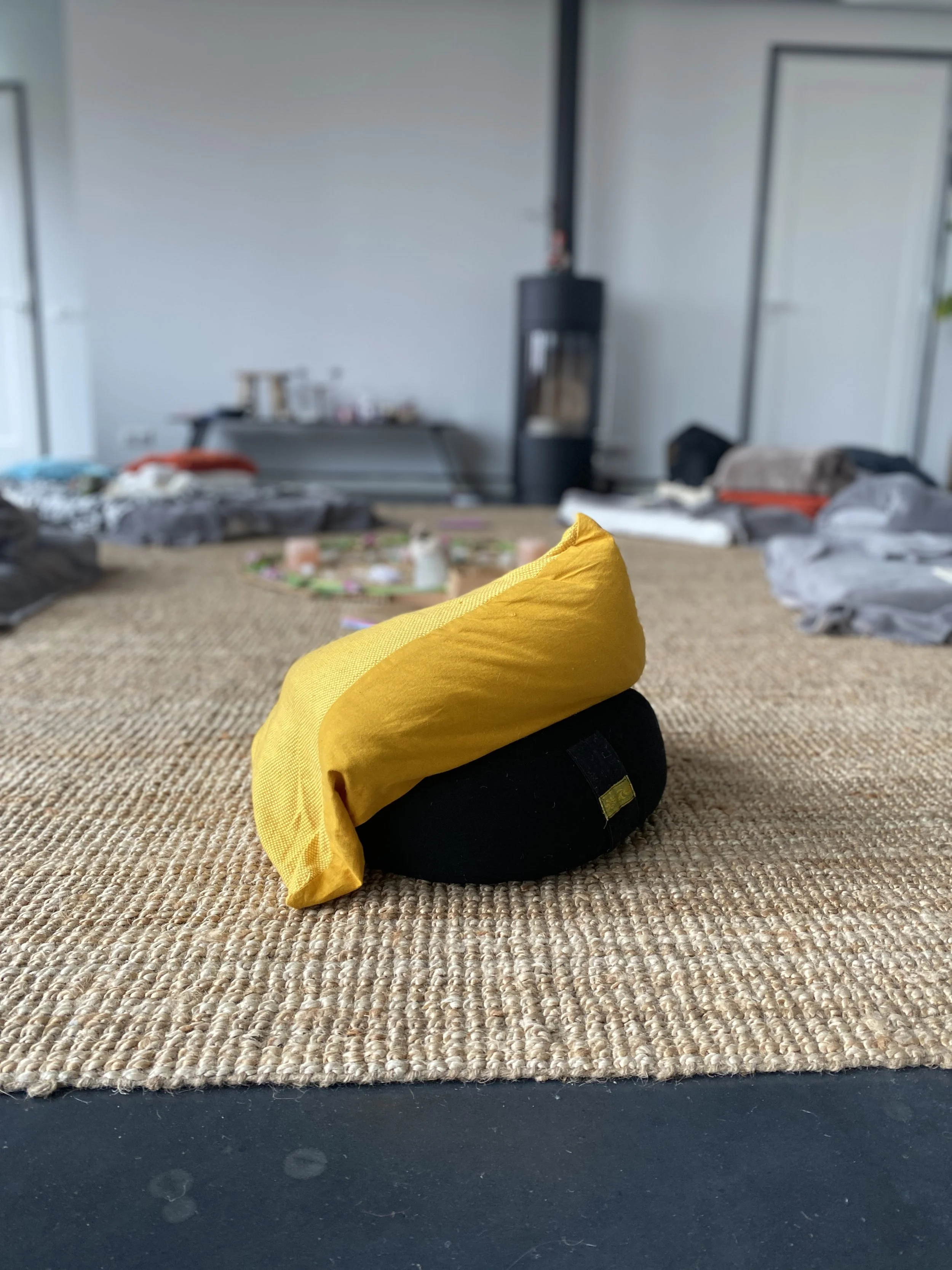 A meditation cushion with a yellow cover on top of a black cushion, placed on a woven rug in a cluttered room with beds and furniture in the background.