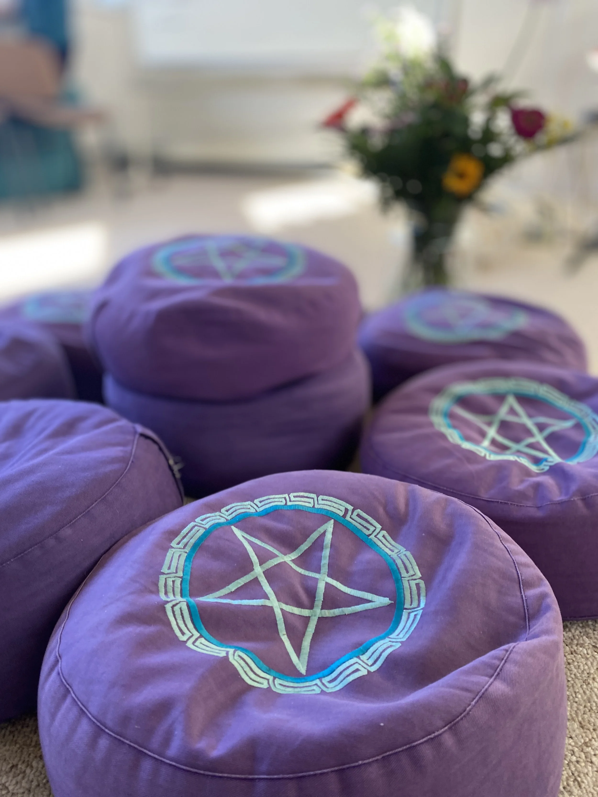 Several purple cushions with a pentagram symbol embroidered on top, placed on a beige carpet near a blurred background with a flower bouquet and a window.