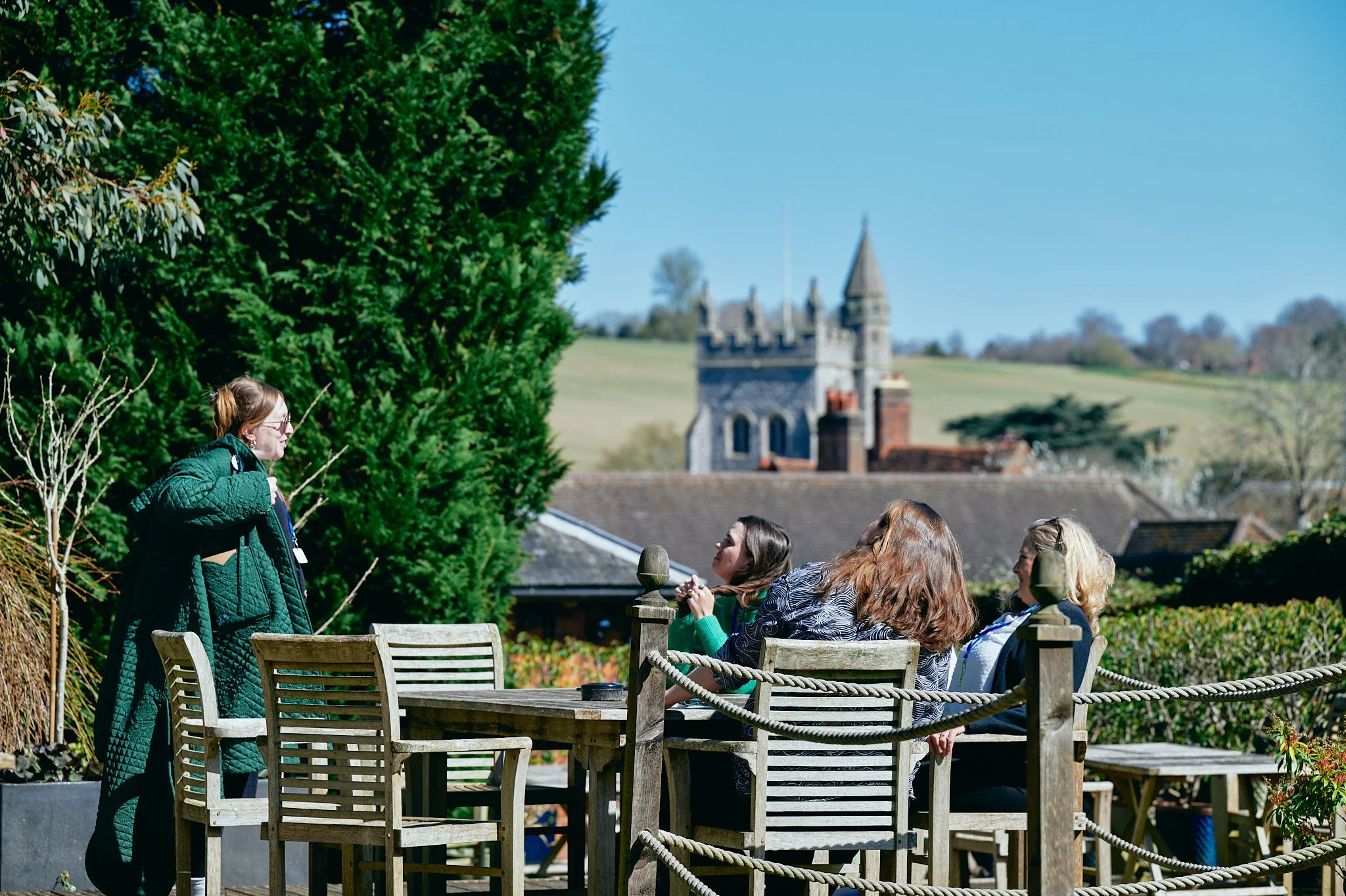 A group of women sitting around a garden table outside, overlooking a village with a prominent church tower.