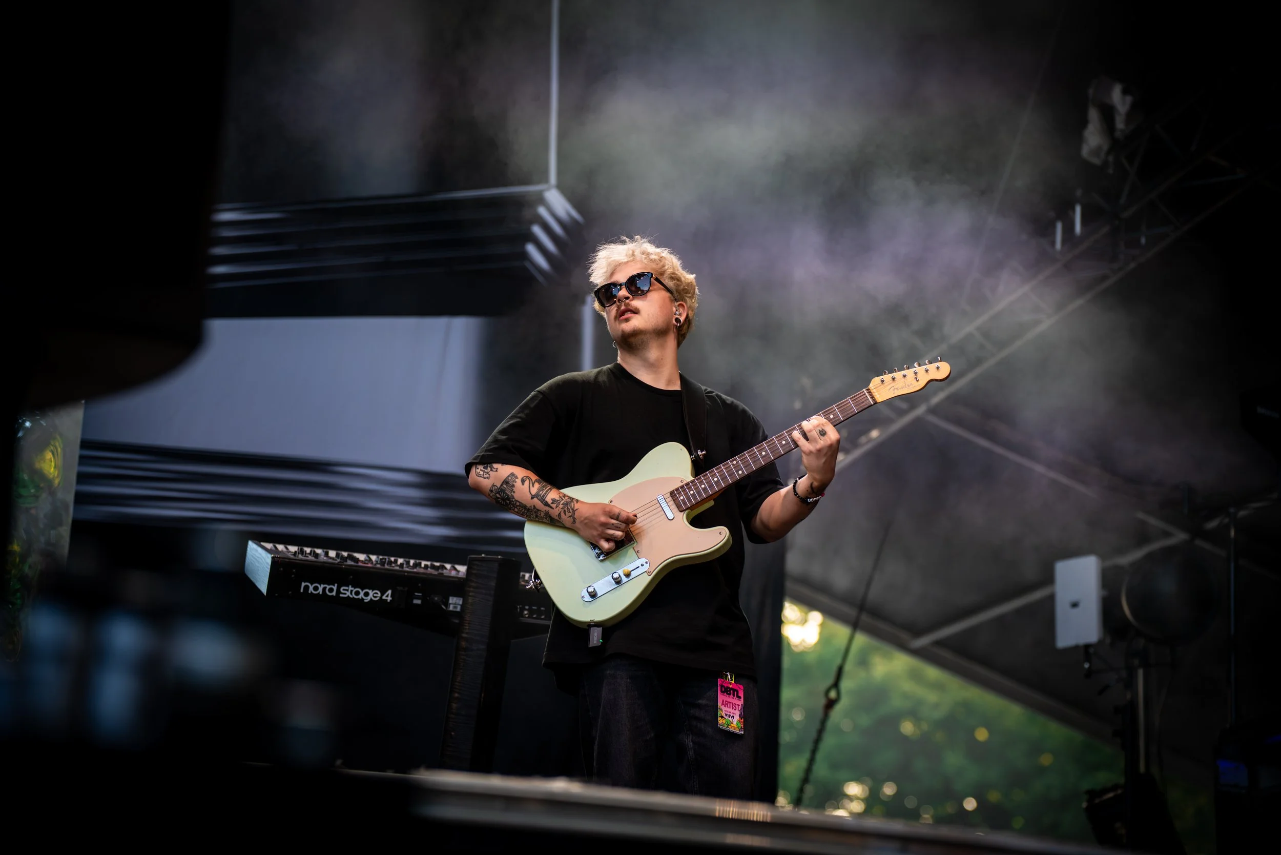 Mies soittaa kitaraa konserttilavalla, taustalla keikkanäyttö ja puupaneelit, päällä tummat vaatteet, aurinkolasit, värilliset lyhythiukset.