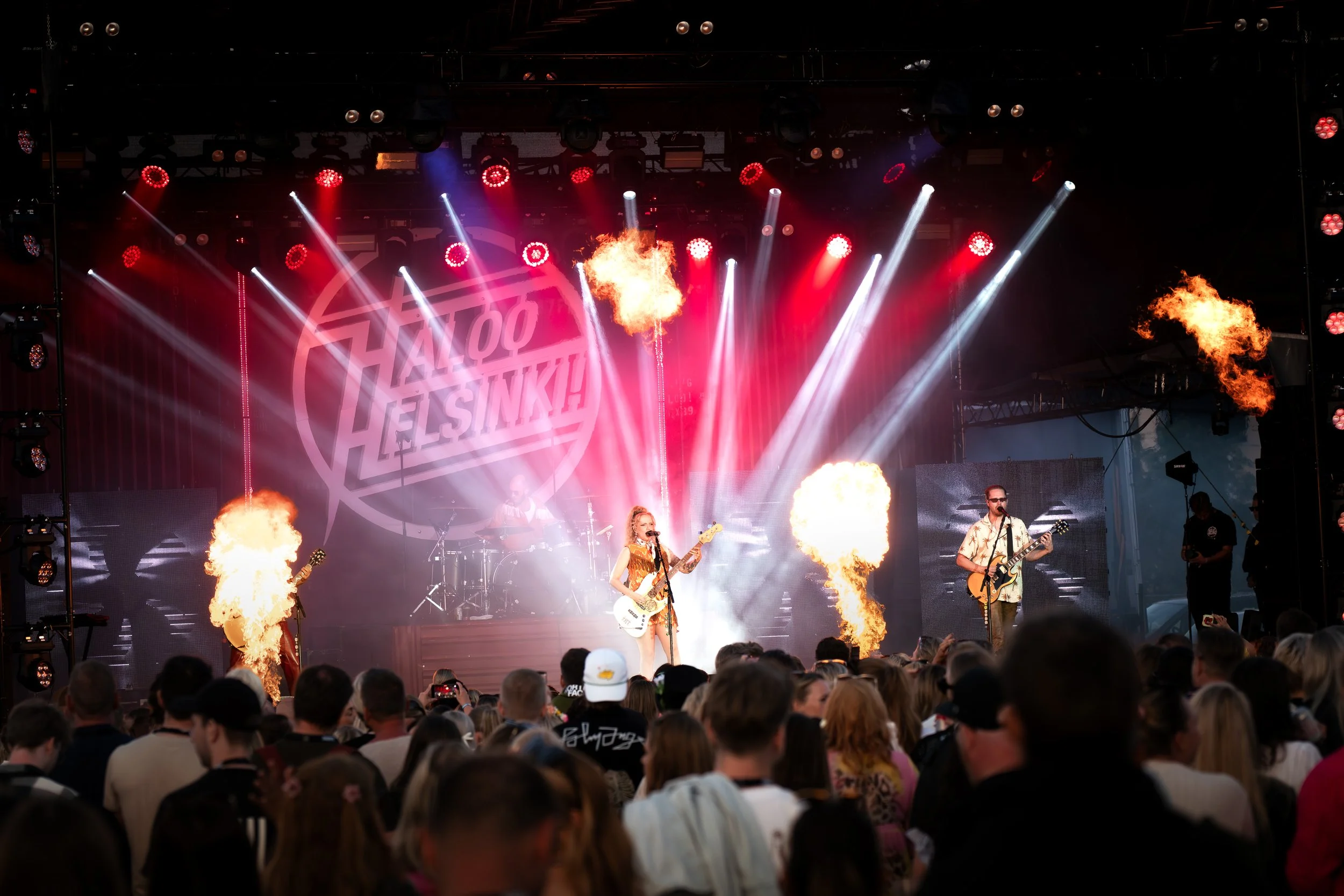 Live rock concert on stage with flames, bright lights, and band performing, audience watching.
