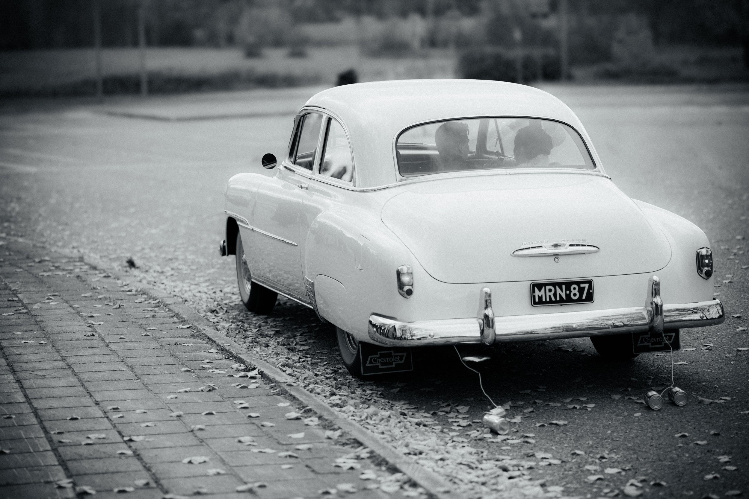 Musta-valkoinen kuva vanhasta autoilma, parkkipaikalla, syksyllä lehdet maassa, kaksi lasten lelulaatikolla auton takana.