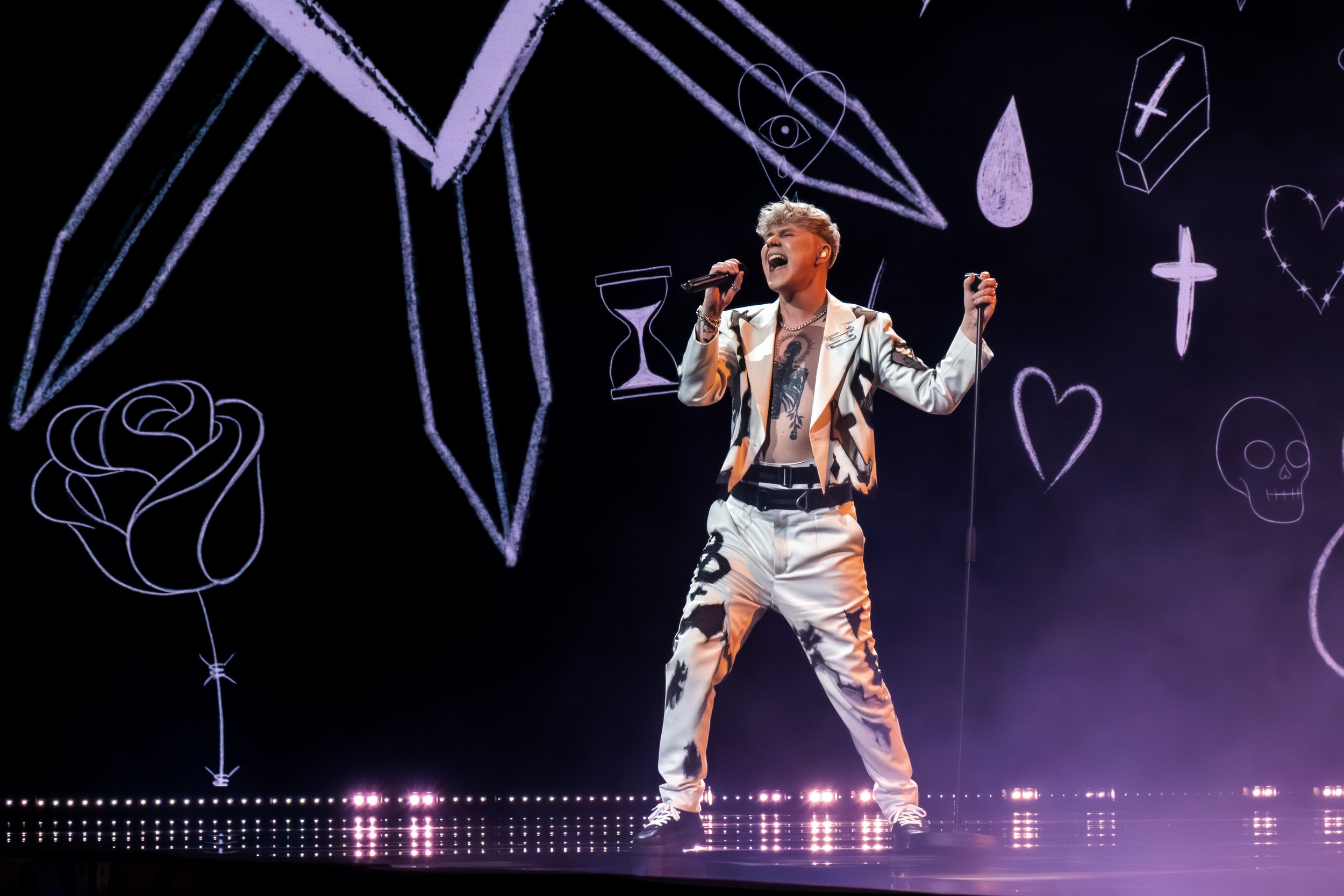 Laulaja esiintyy lavalla, taustalla on sateenkaaren väreissä piirrettyjä symboleja kuten sydän, kallo, risti ja kynä, ja hän käyttää valkoista ja mustaa asua.