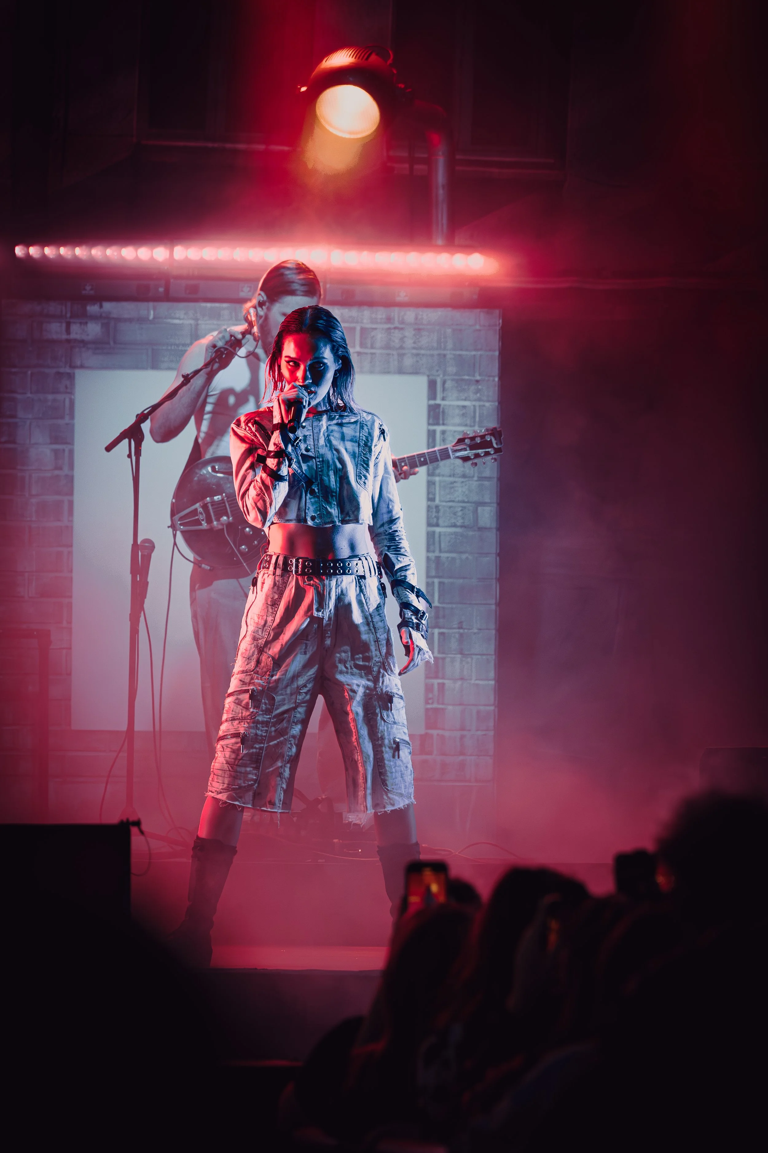 Nuori nainen laulamassa mikrofonilla lavalla, taustalla kitara soittaja, punaisia ja sinisiä valoja, yleisöä etualalla.