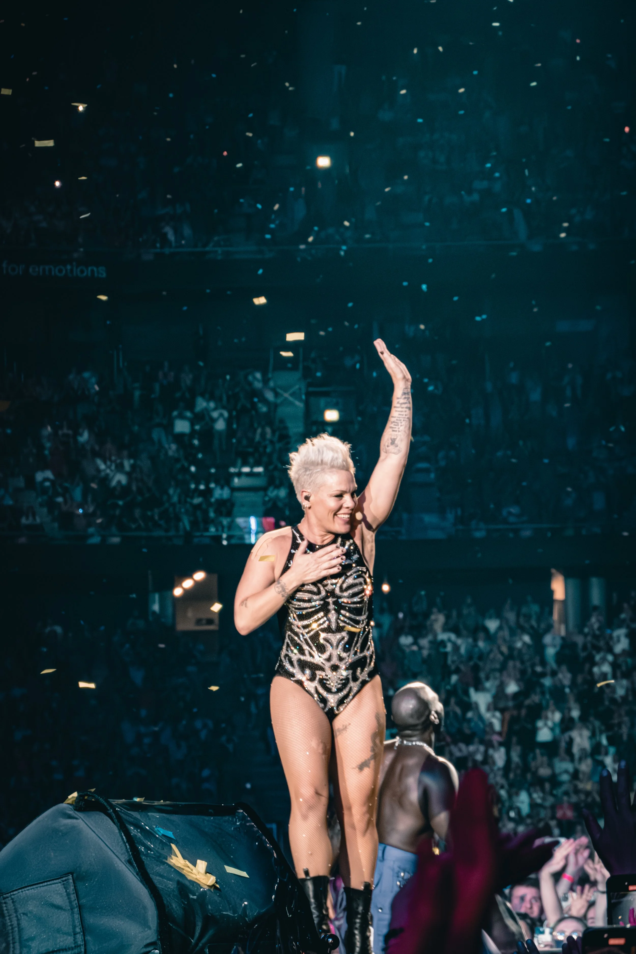 Performer on stage in a crowded concert hall, wearing a black and gold outfit, with one hand on her chest and the other raised in the air.