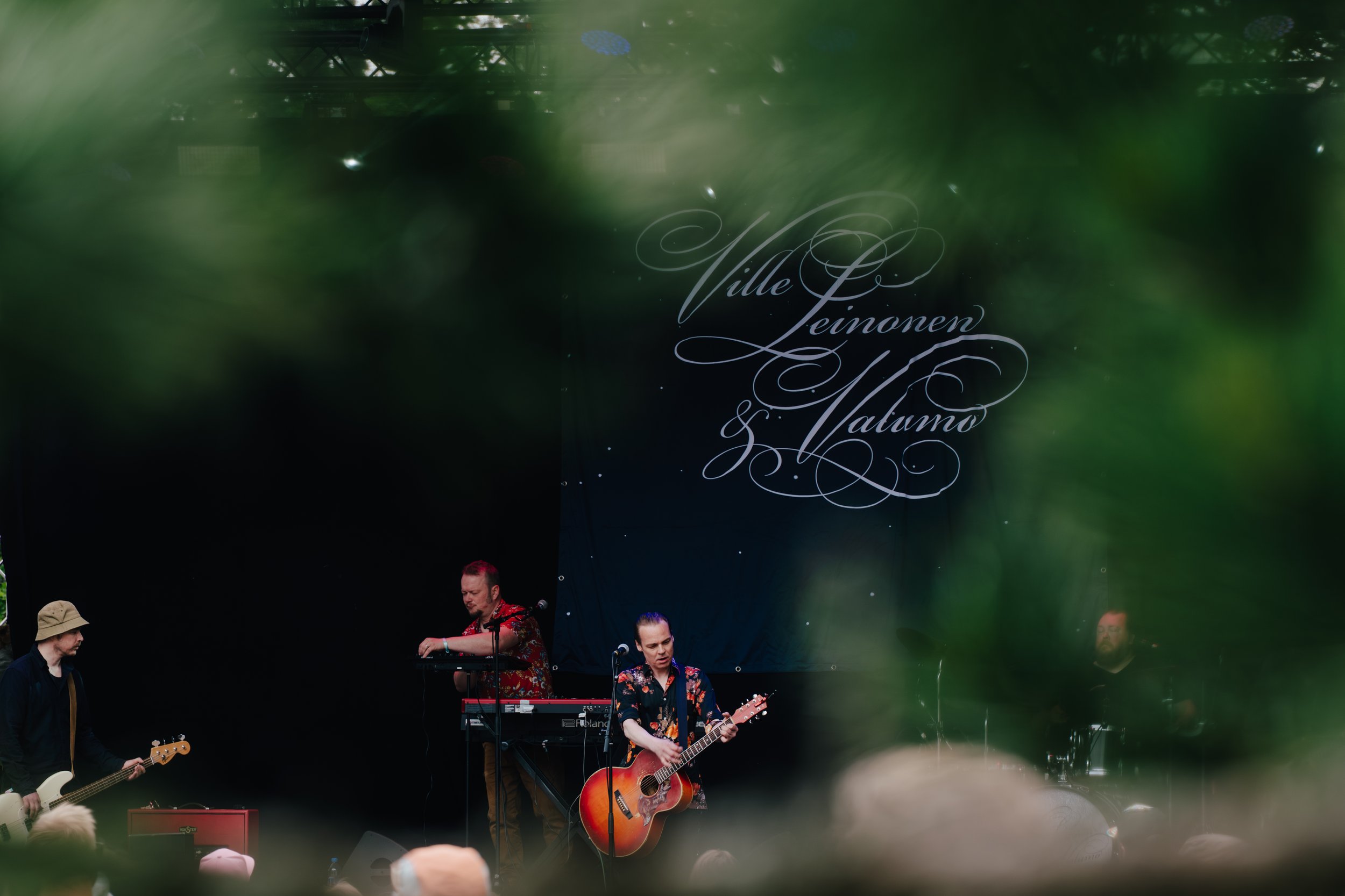 Kolmimies soittaa kitaraa ja laulaa, toinen soittaa koskettimia ja toinen sivusilmällä katsoo, musiikkiesityksessä, taustalla teksti 'Ville Leinonen & Valumo', yleisöä edessä