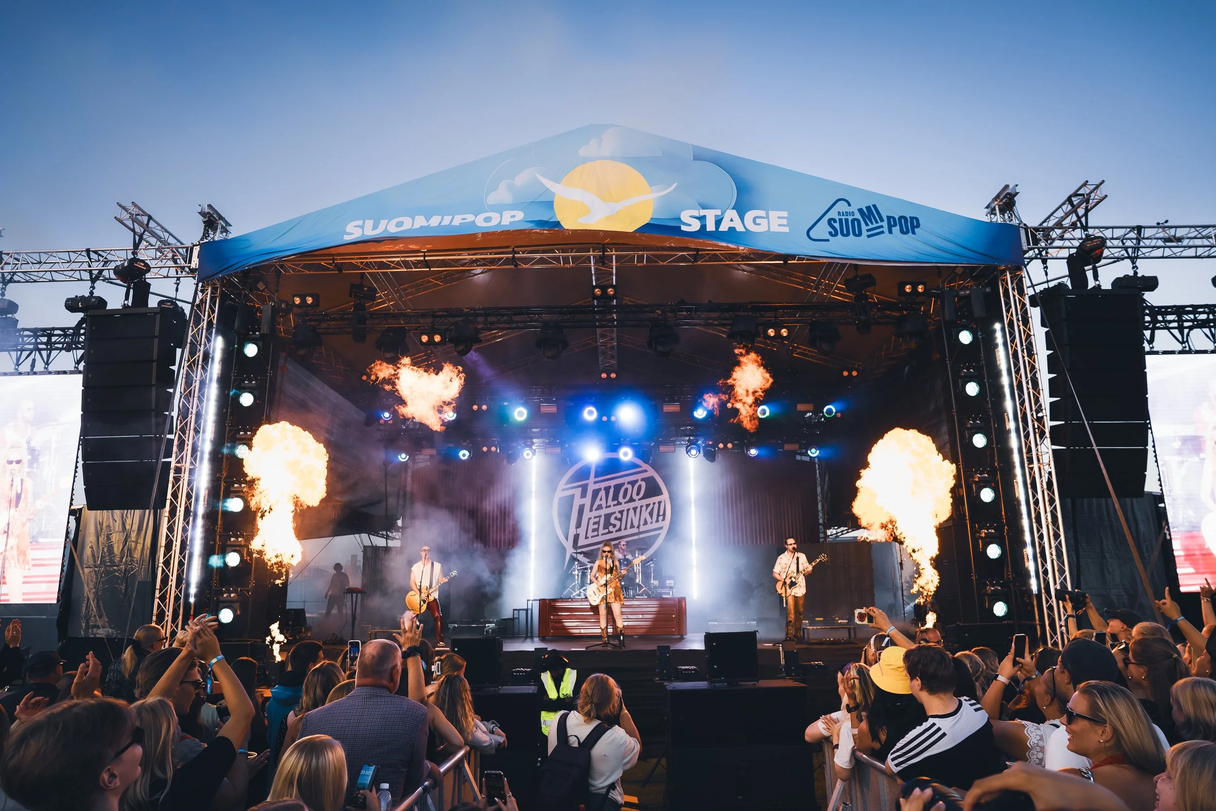 Elävän musiikin konsertti ulkoilmassa, suuri yleisö ja lava, jossa bändi ja tuliefektit