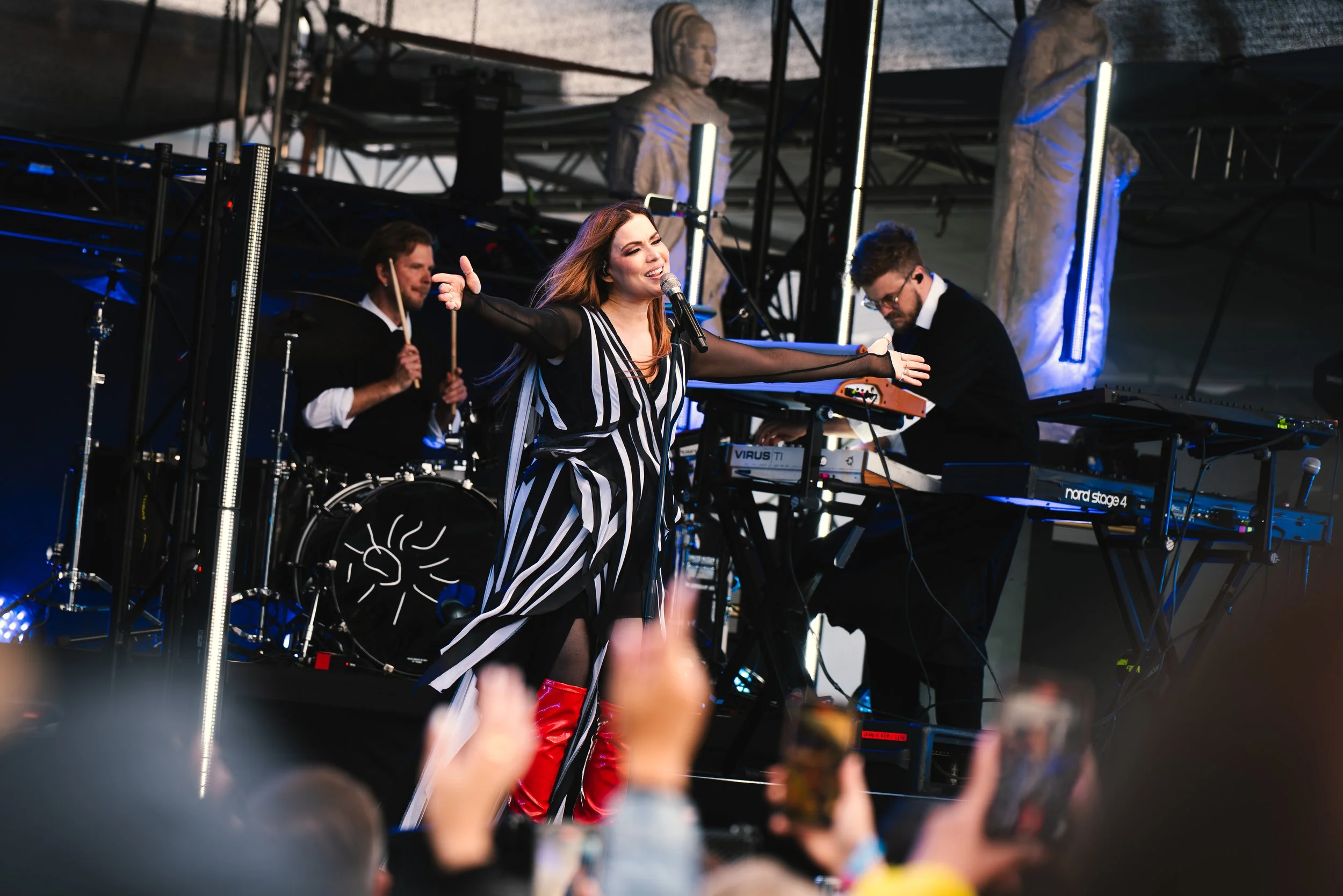 Nainen esiintyy lavalla bändin kanssa, laulaa ja soittaa koskettimia, taustalla rumpali ja toisen koskettimien soittaja.