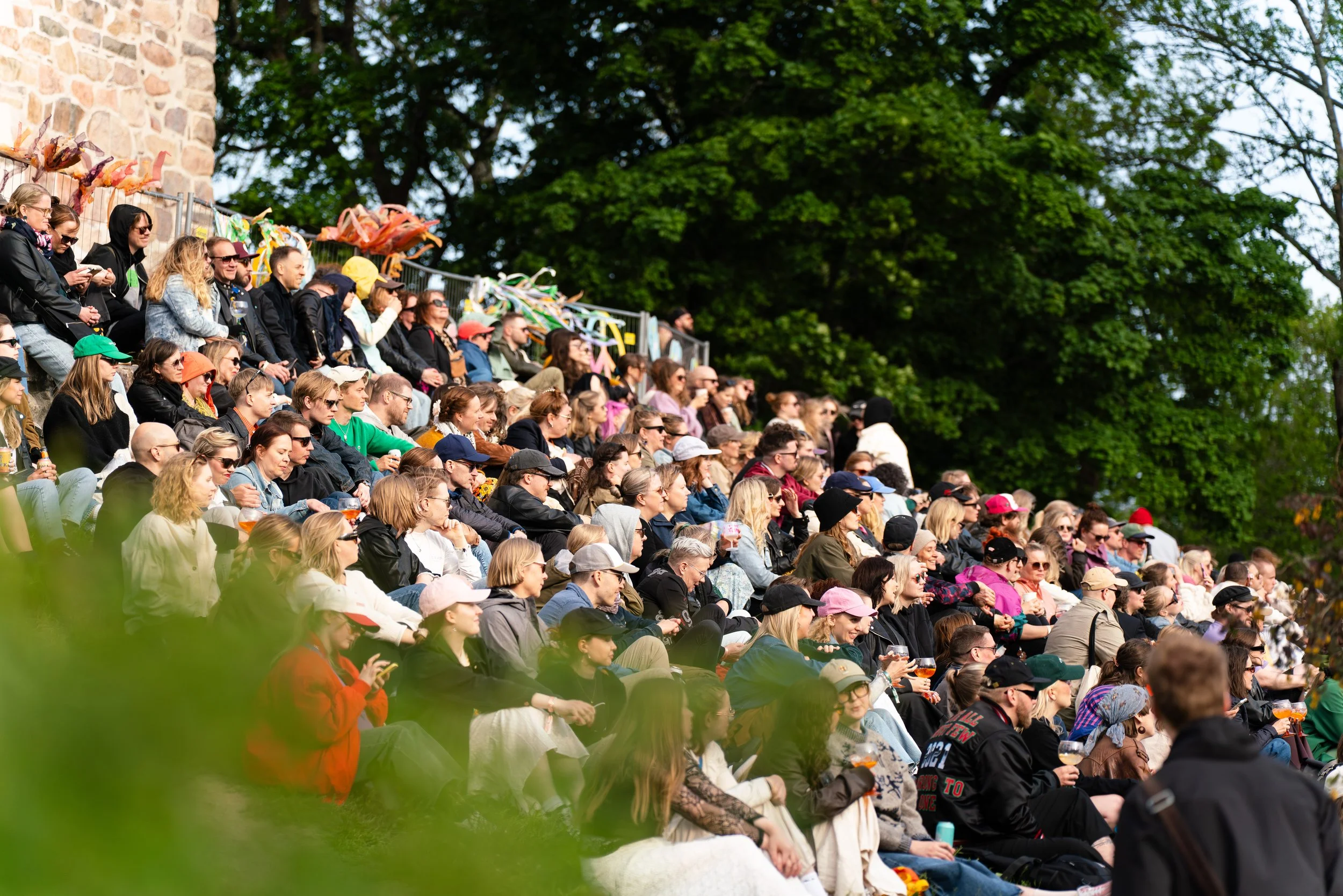 Ihmisjoukko istuu katsomossa katsomassa ulkoilmatapahtumaa, taustalla puita ja osa rakennuksesta, kesäinen sää