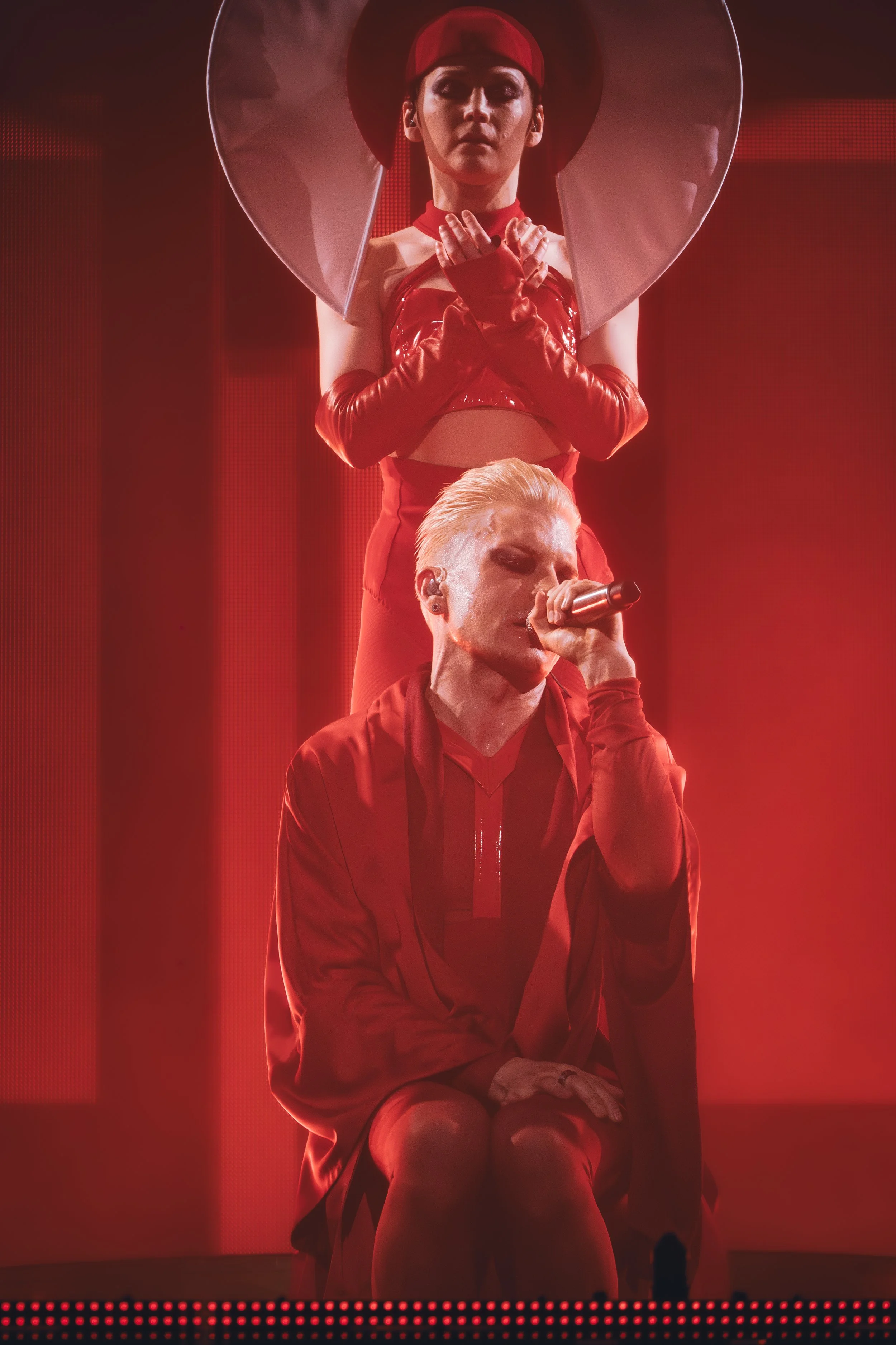 Kaksi henkilöä esiintymislavalla, takana nainen silinterihatussa ja punaisissa ja takana mies istuen, puhuen mikrofonilla, kaikki punaisessa valossa.