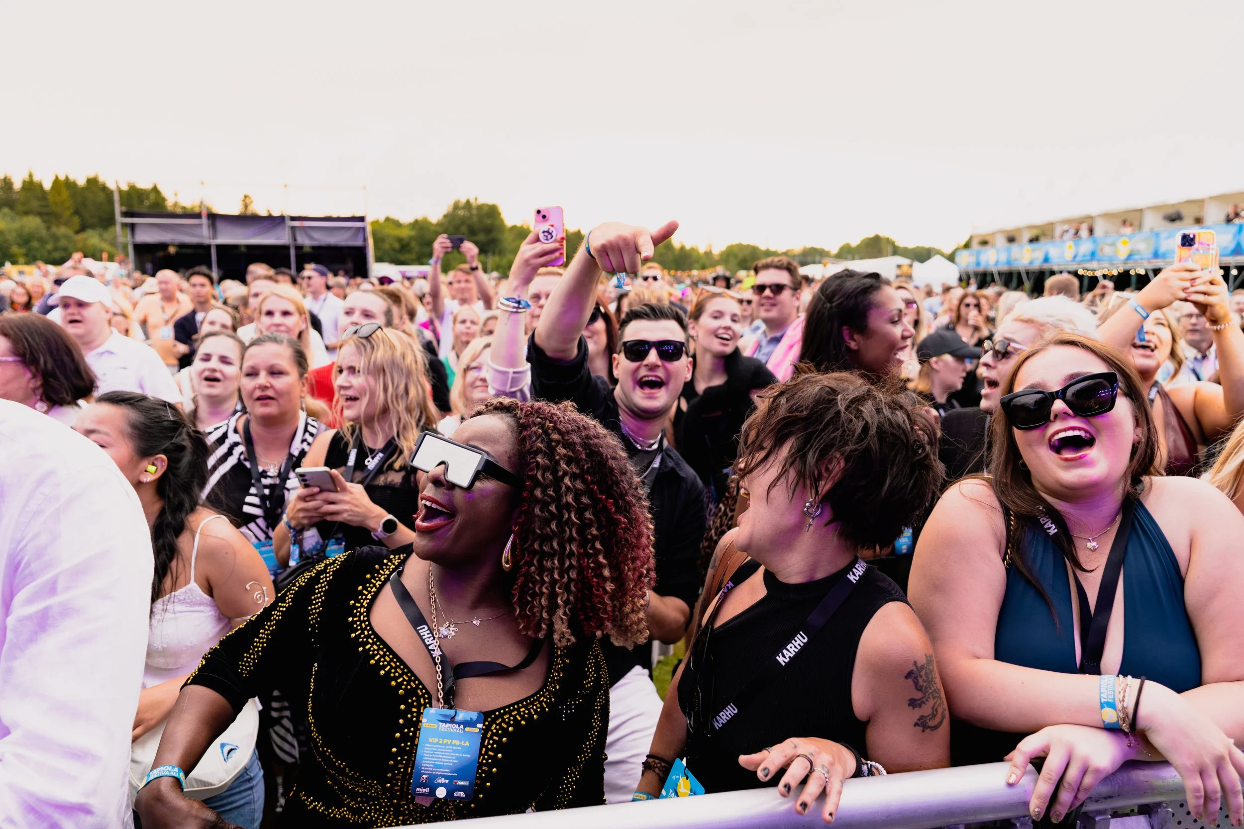 Iloisia ihmisiä nauttimassa musiikkifestivaalista, osa ihmisistä käyttää aurinkolaseja ja selfie-kameroita, olemassa suuria yleisöjä ulkona konserttialueella.