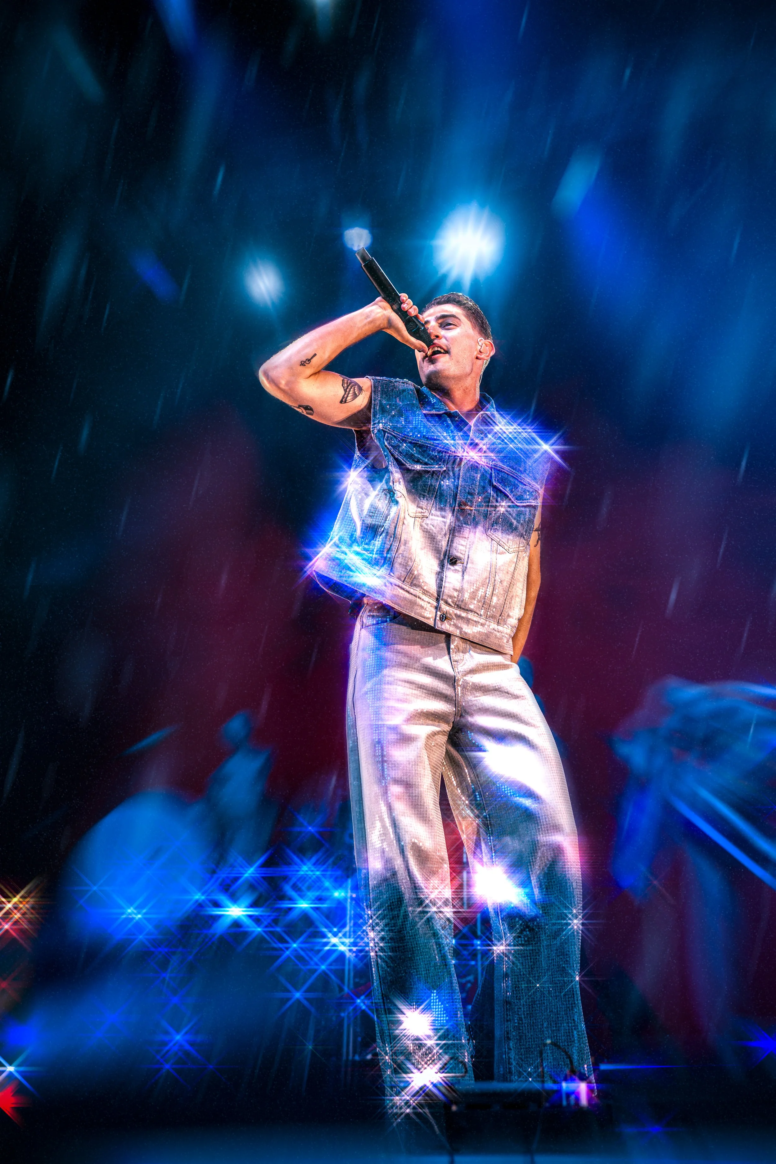 Xkmäzetu musiikkiesiintyjä runsaan valon ja värikkäiden efektien keskellä, pitää mikrofonia kädessään lavalla.