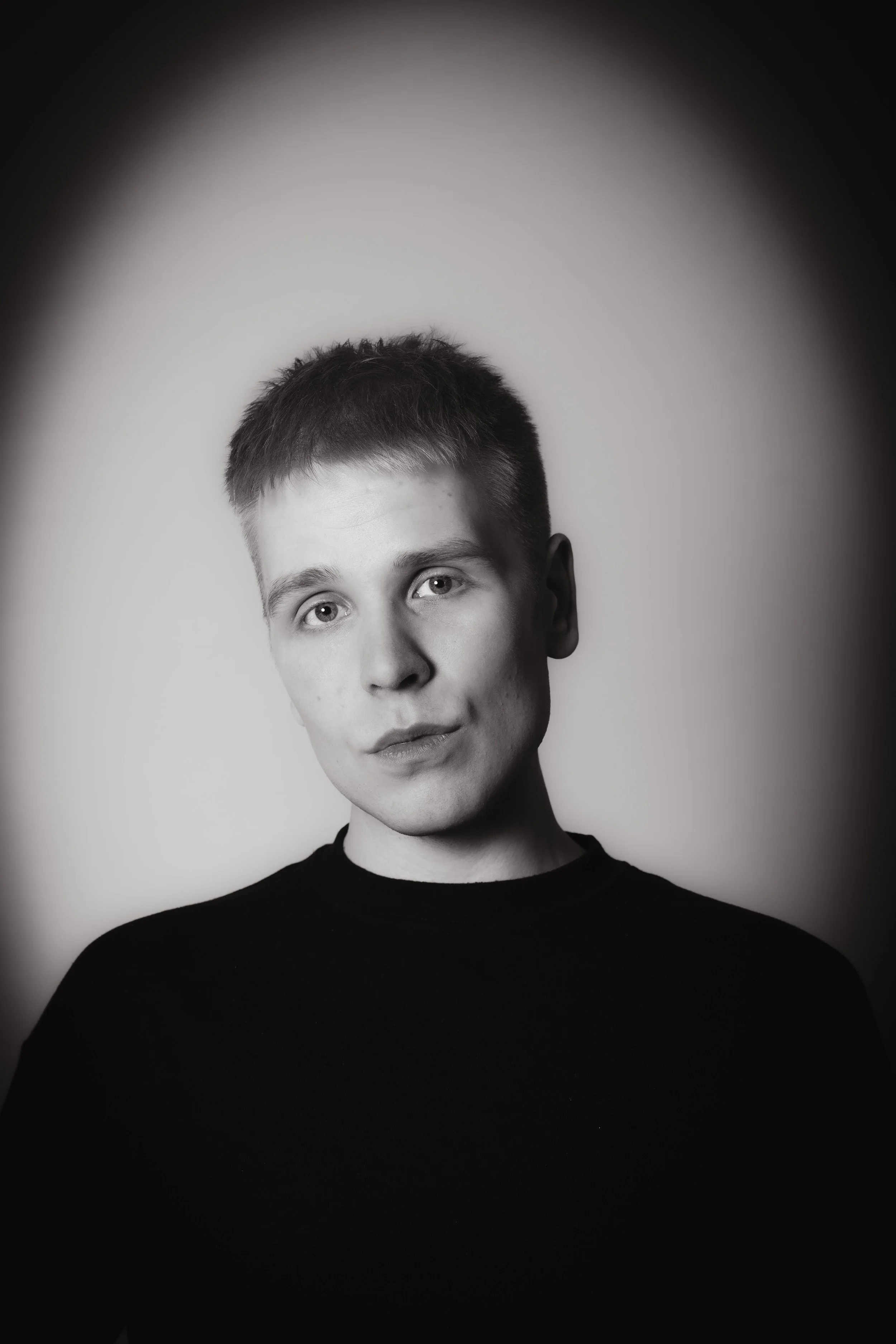 Mustavalkoinen valokuva nuoresta miehestä, jolla on lyhyt tukka, katsovat suoraan kameraan neutraalilla ilmeellä. Tausta on vaalea, ja hänen yllään on musta paita, kuvassa on pehmeä valaistus ja tumma kehysneutraali kehyksen reunoja.