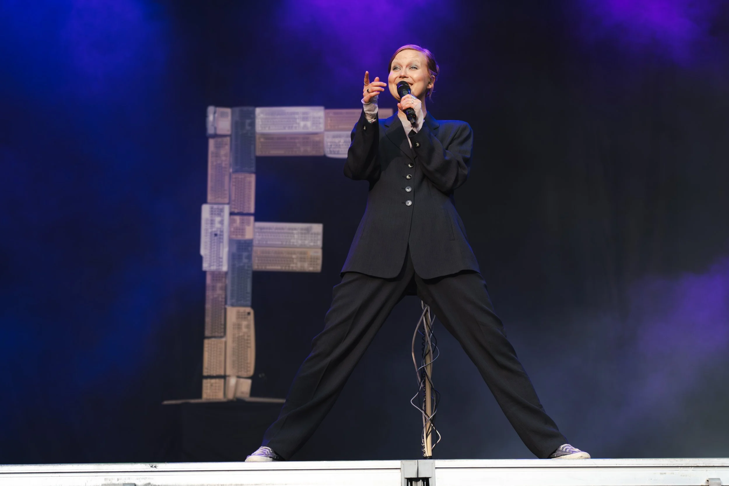 Nainen esiintyy lavalla, pitää mikrofonia ja hymyilee, taustalla kirjaaiheinen taidetyö.