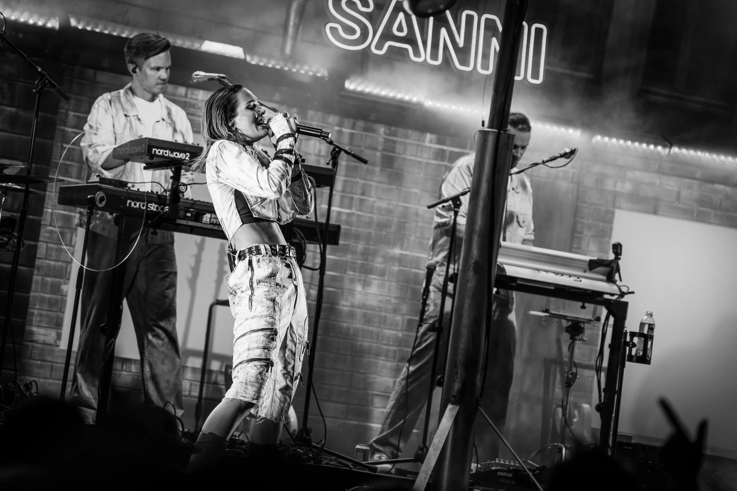 Nainen esiintyy laulamalla mikrofonilla, taustalla kaksi miestä soittamassa syntetisaattoreita, esiintymispaikka nimeltä 'SANNI' näkyy taustalla.