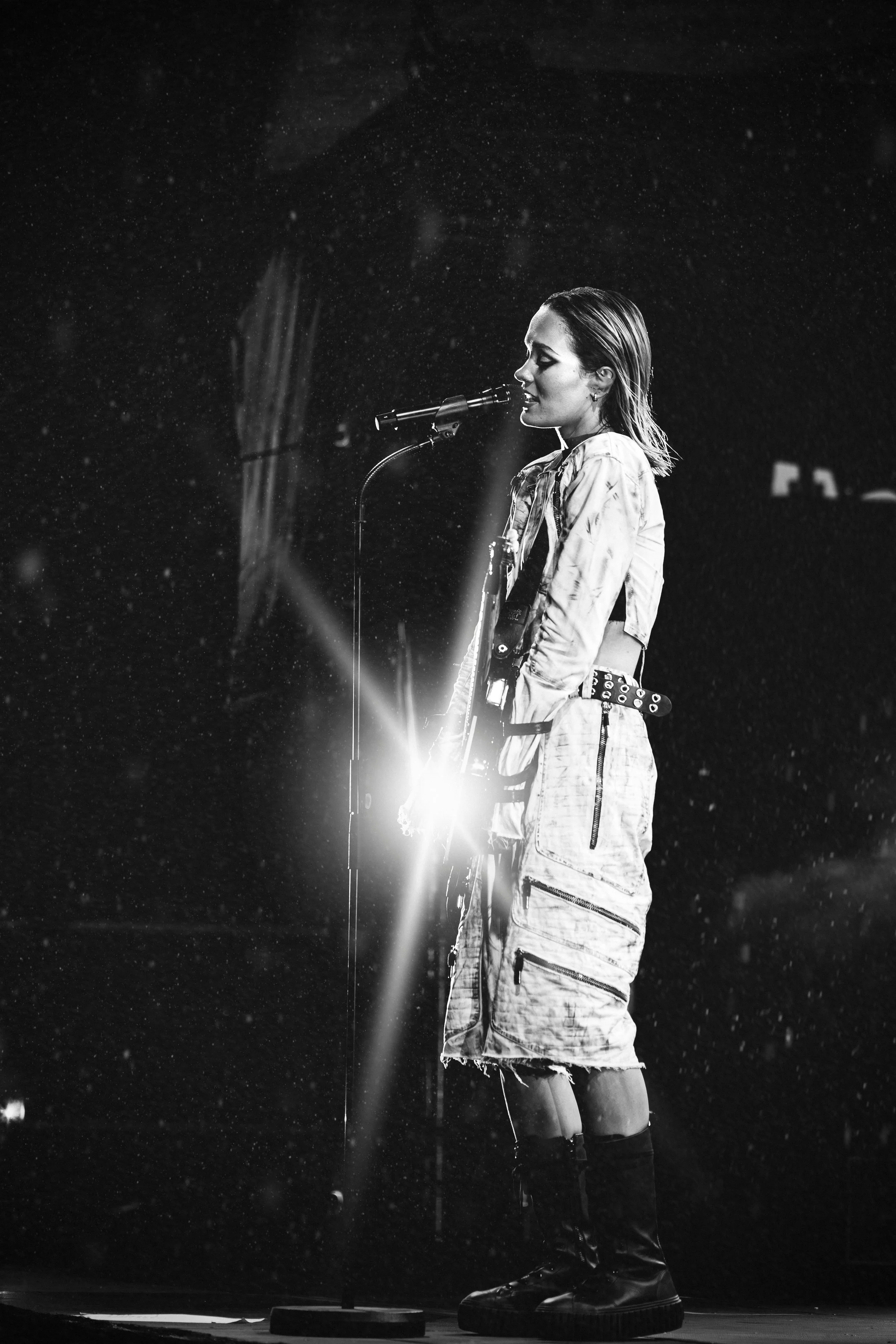 Mustavalkovalokuva naisesta laulamassa mikrofonin edessä, taustalla ja ympärillä valon ja sateen vaikutelma.