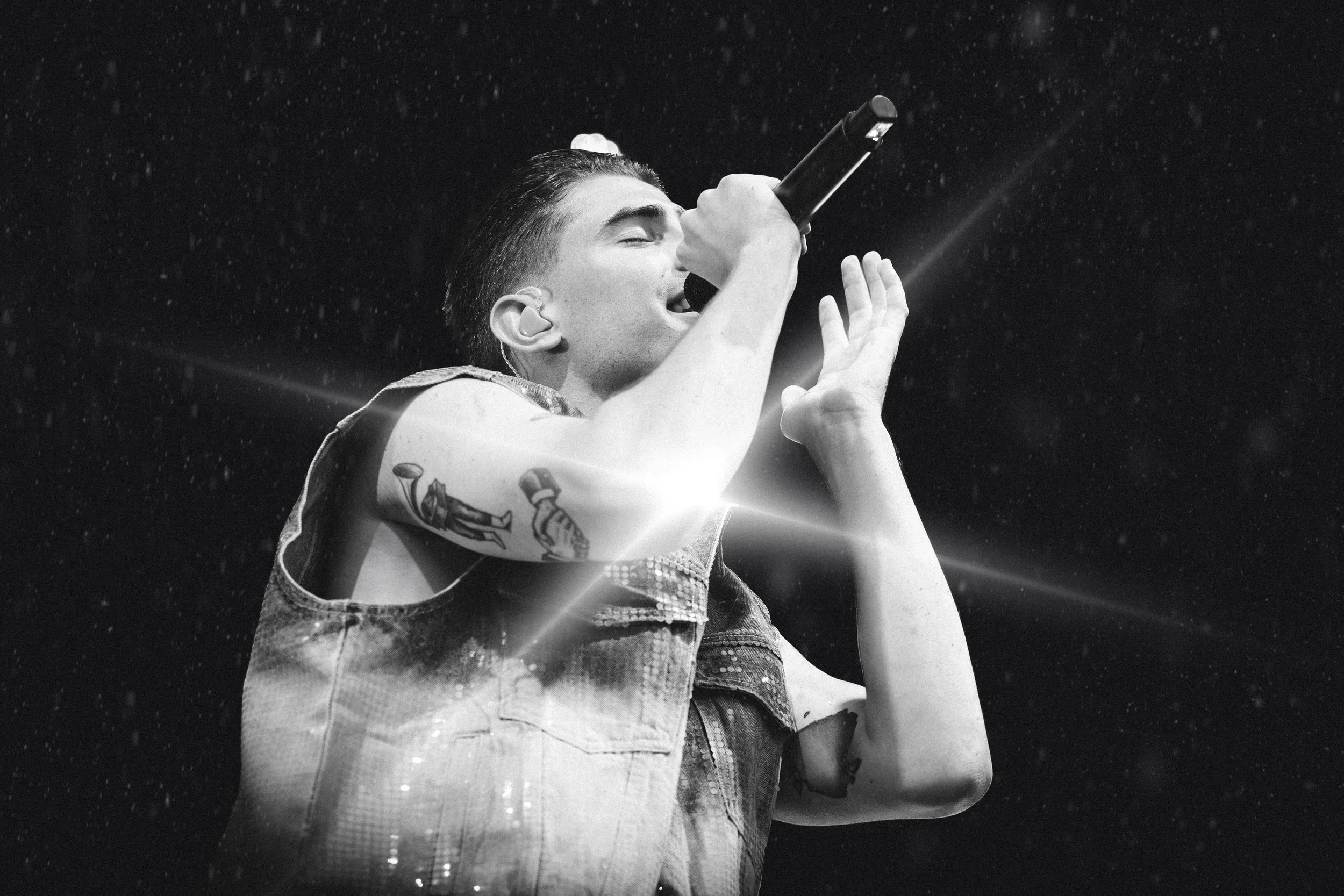 Mies esiintyy mikrofonin kanssa mustavalkokuvassa, taustalla pieniä vesipisaroita ja valojuovia.