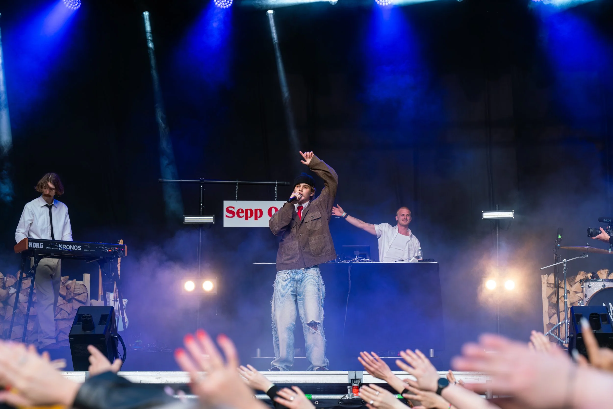 Bändi esiintyy lavalla, keskellä mies laulaa mikrofonilla, taustalla soitin soittaa ja DJ:llä on tietokone. Yleisöä on etualalla, osan käsistä näytetään.