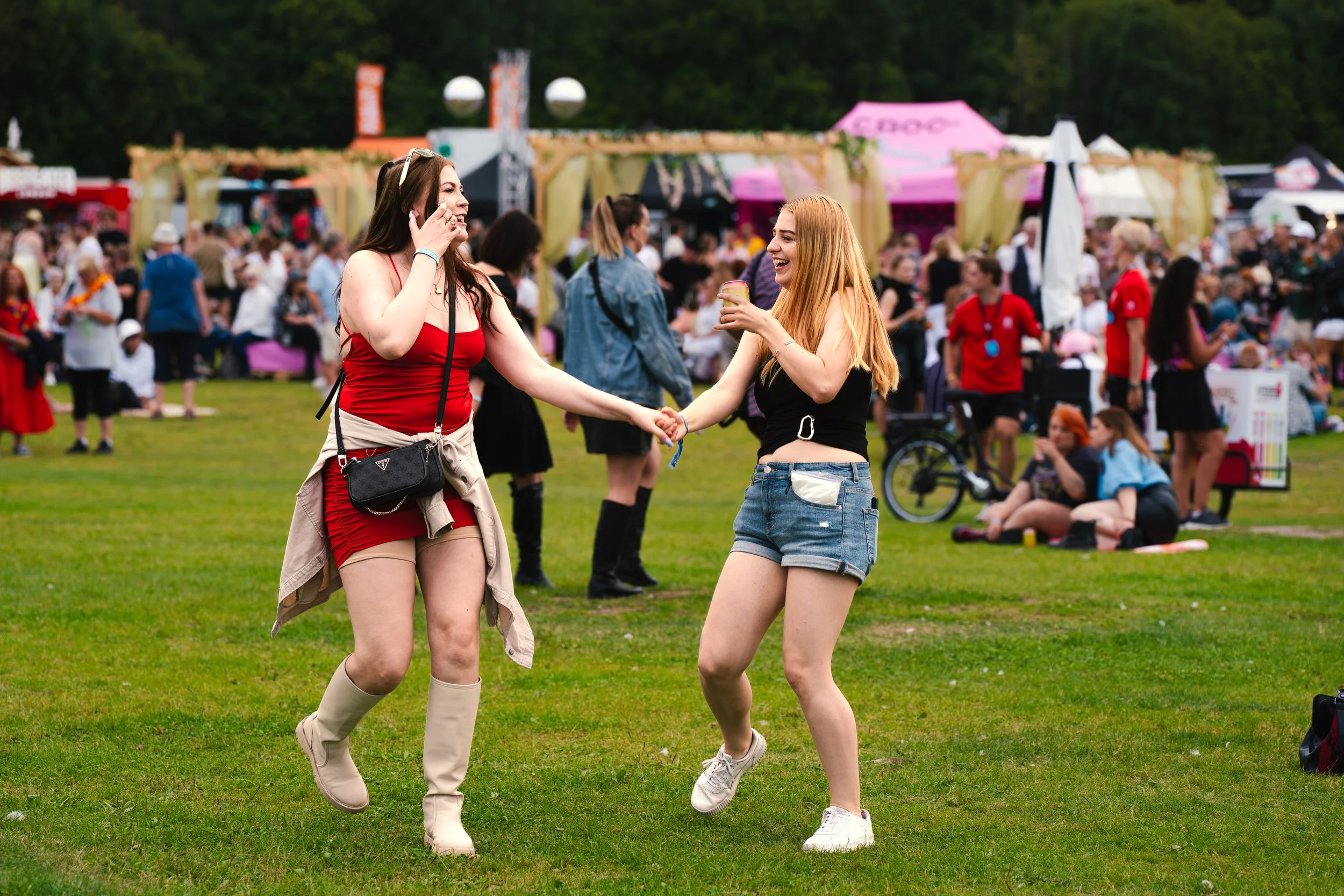 Kaksi nuorta naista tanssii käsi kädessä festivaalialueella, taustalla muita ihmisiä ja festivaalikojuja.