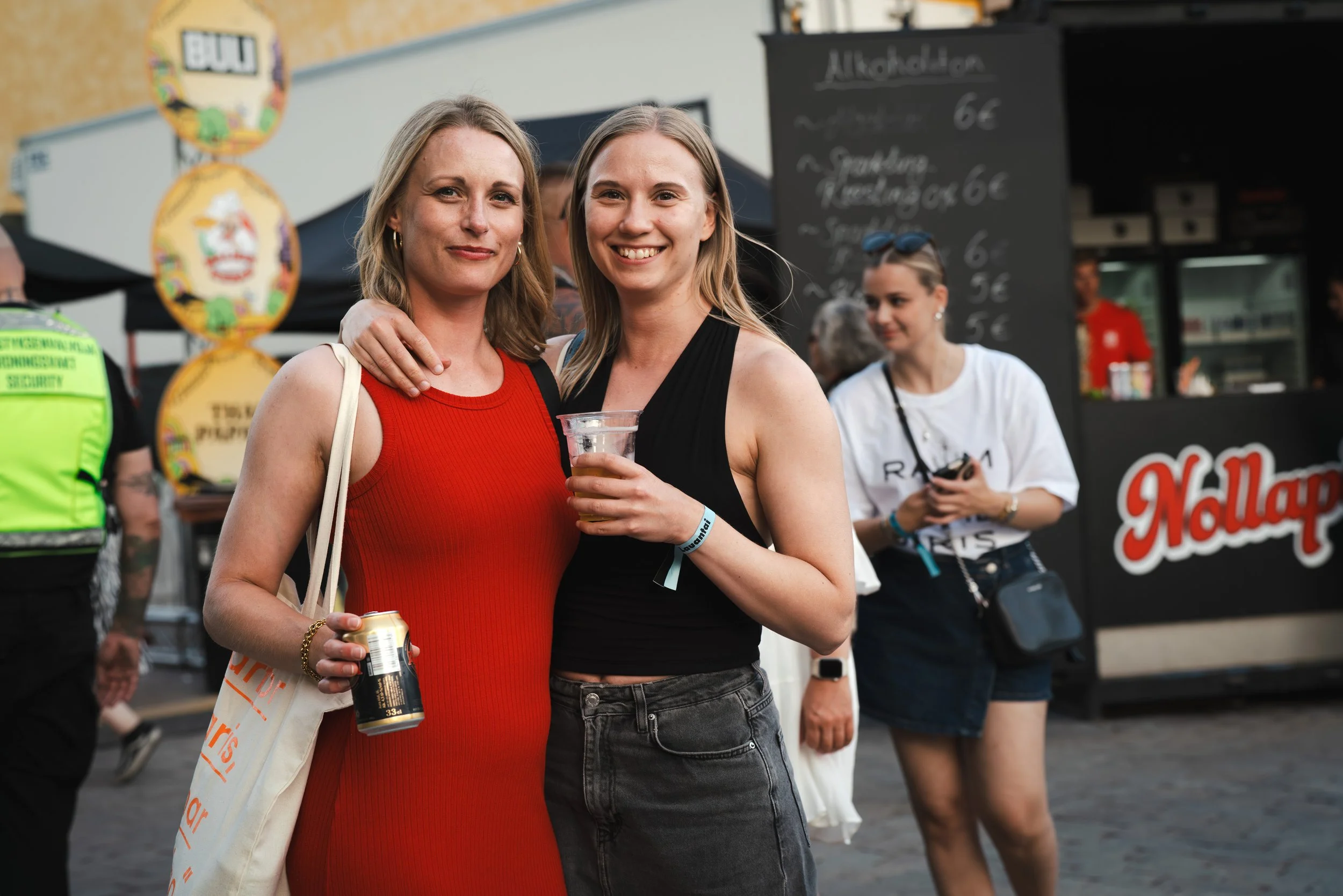 Kaksi nuorta naista hymyilevät ja ottavat selfie-kuvaa festivaalialueella, toisella heistä on lasi olutta ja toisella tölkki juomaa, taustalla ruokakoju ja muita ihmisiä.