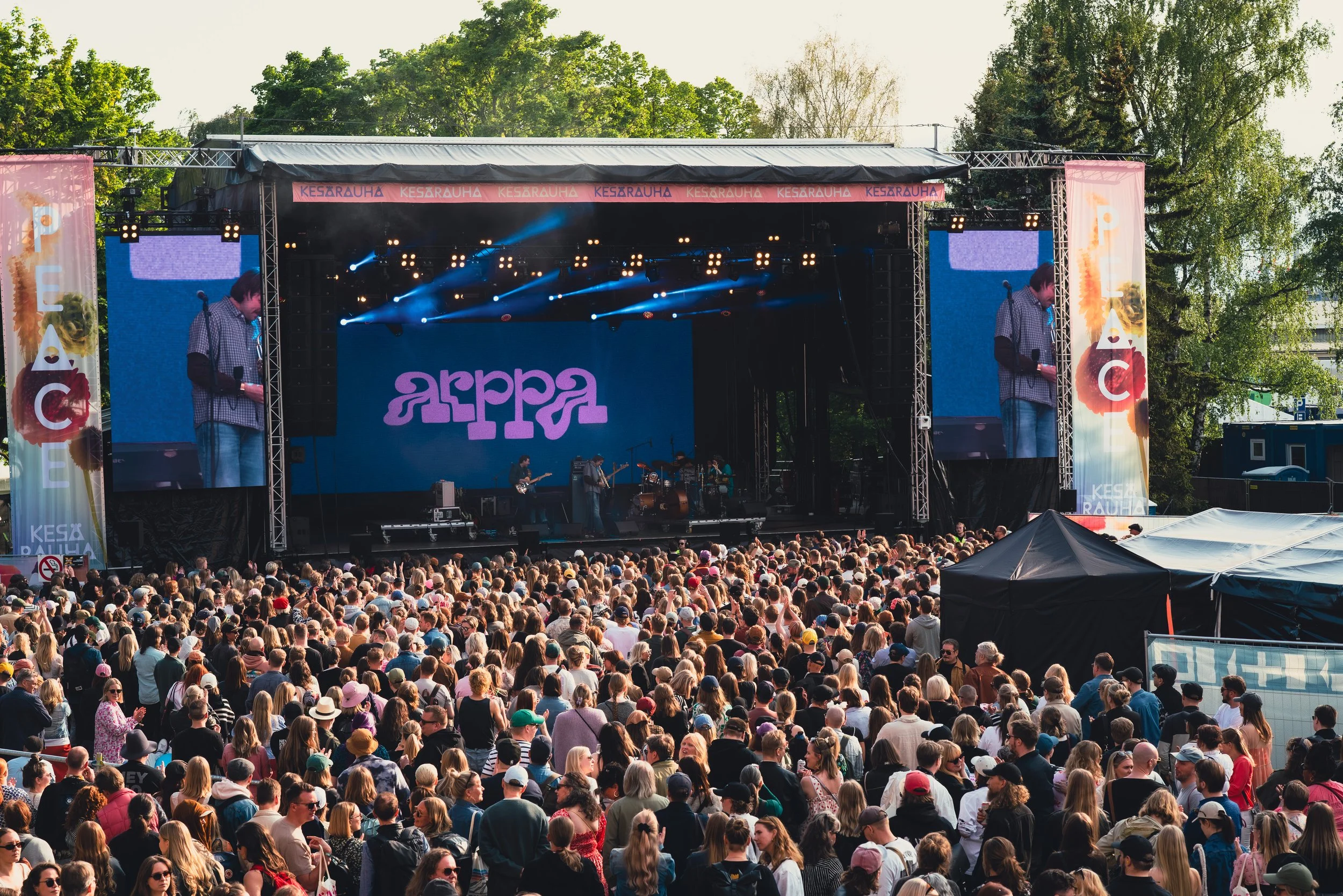 Suuri ihmisten yleisö konserttitapahtumassa ulkona, esiintyjä ja bändi lavalla, monikameranäkymä.