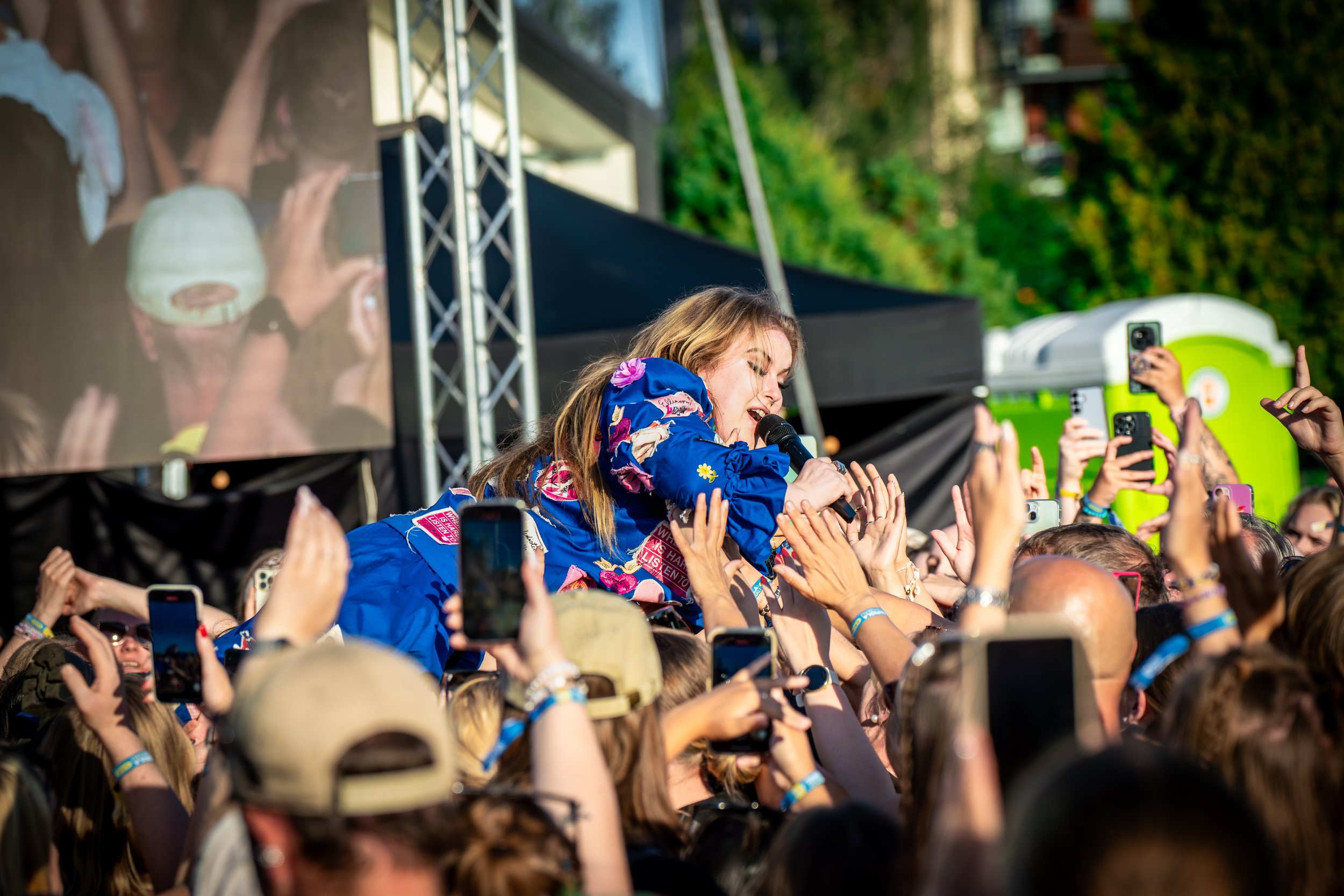 Naistarreinen naisartisti esiintyy yleisön edessä ulkoilmatapahtumassa, hymyilee ja pitää mikrofonia, ympärillä paljon ihmisiä, jotka kuvaavat tapahtumaa puhelimillaan.
