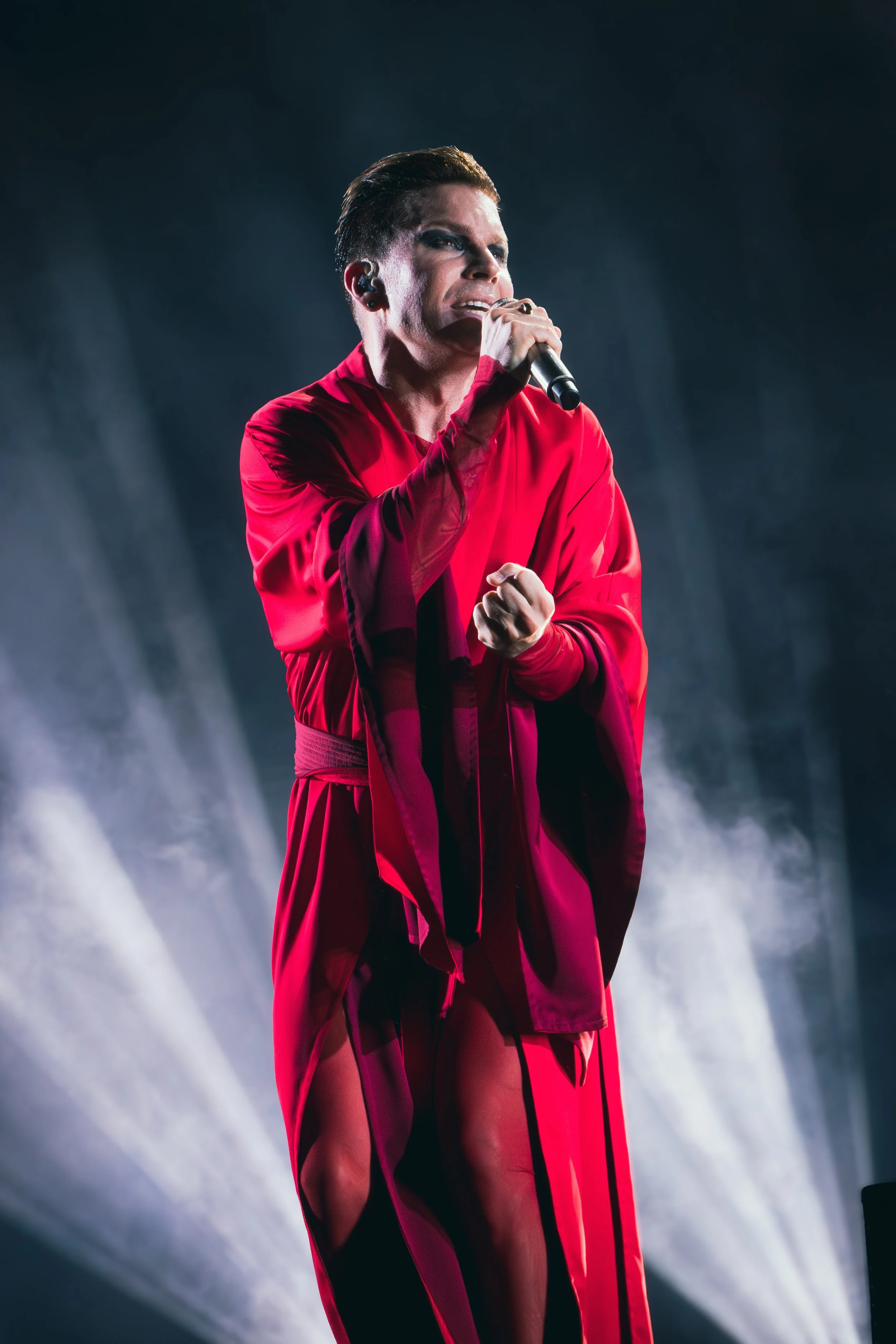 Vokaali esiintyy lavalla musiikkiesityksessä, pukeutuneena punaisiin vaatteisiin ja käyttämällä mikrofonia.