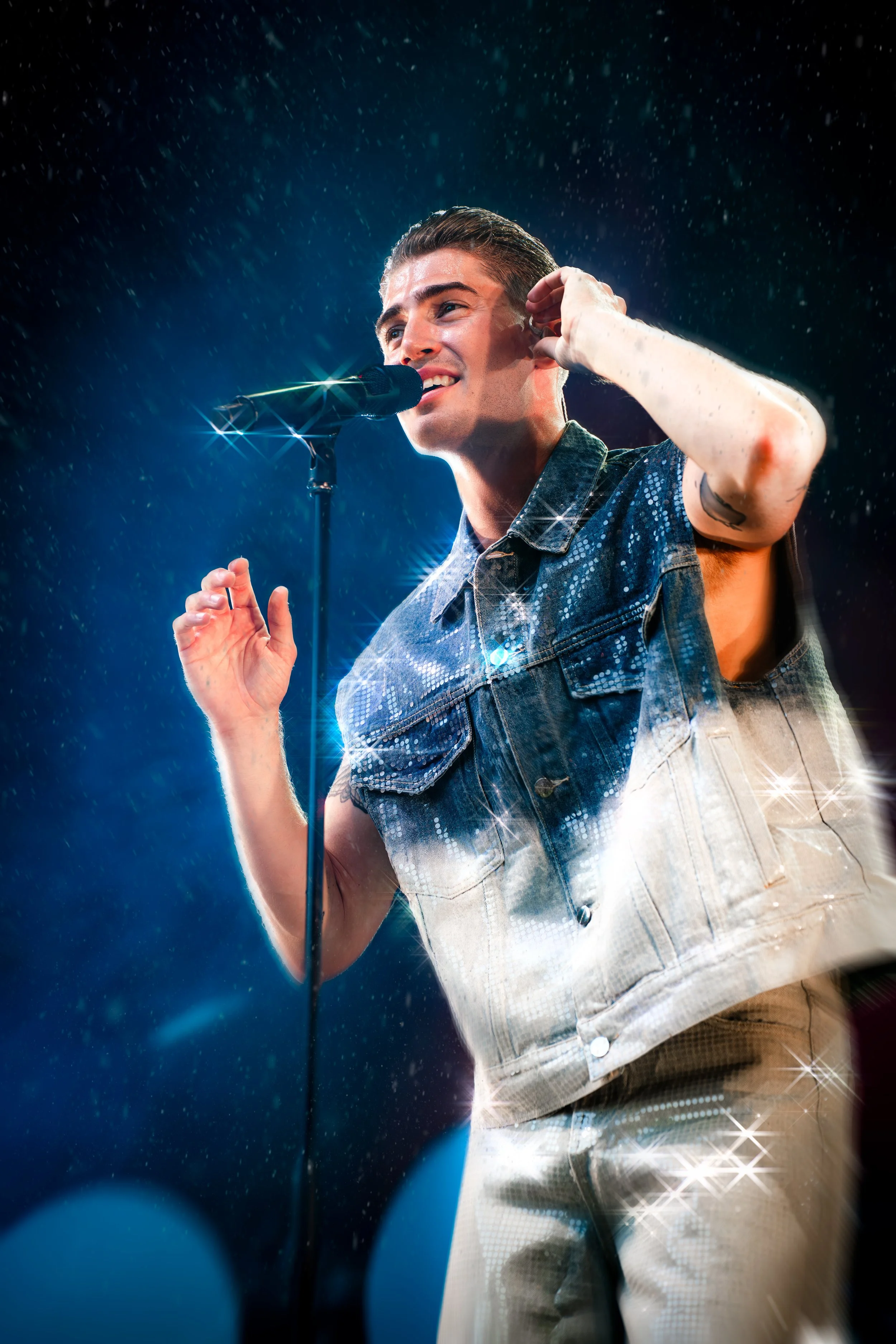 Nuoruusmies esiintyy lavalla mikrofonin ääressä kiiltävässä vaatetuksessa, taustalla sataa vesisateessa ja on sälpää valoja.