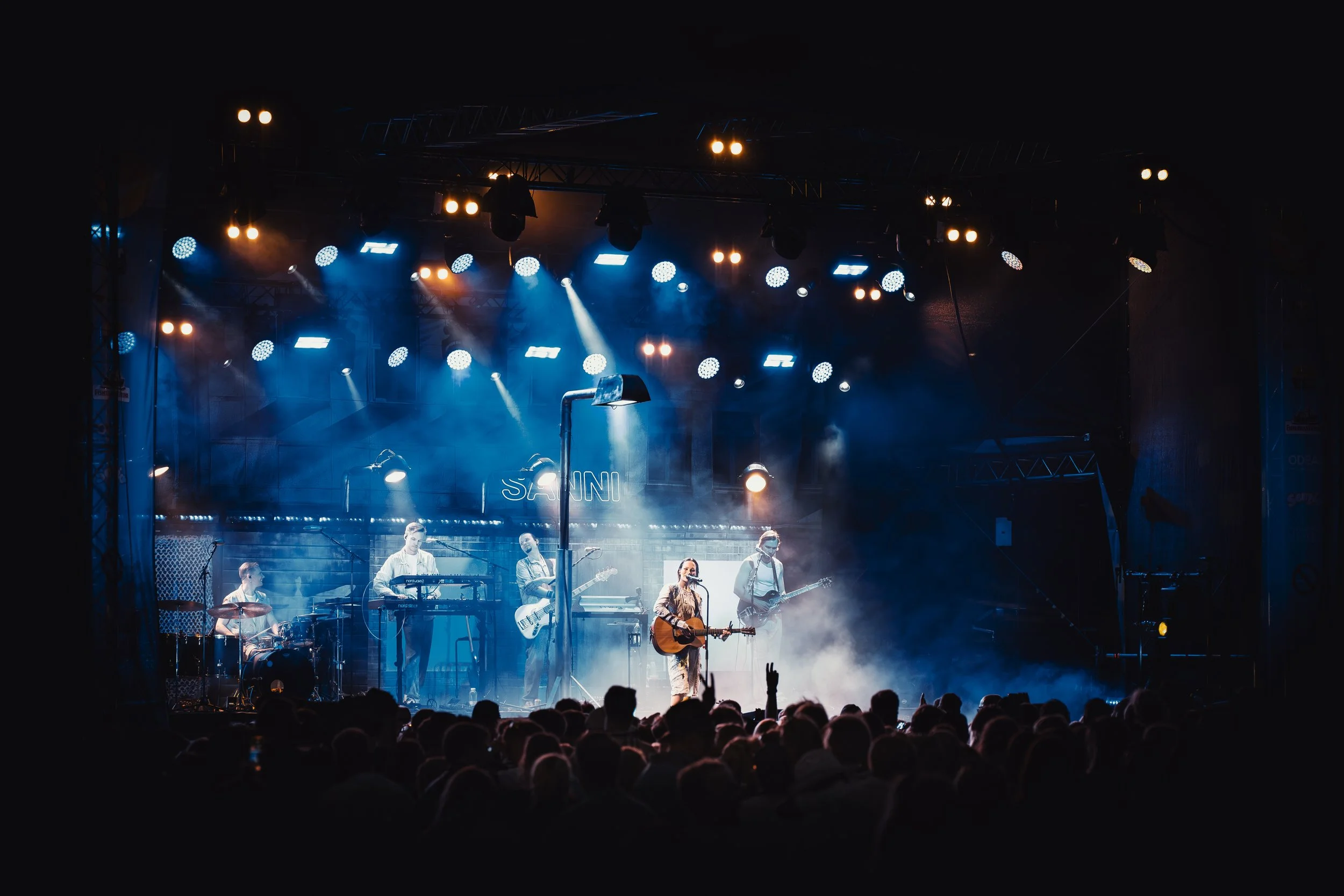 Live musiikkiesitys lavalla, jossa on kuusi muusikkoa ja suuri yleisö edessä. Lattialla ja taustalla on valoefektejä ja savua.