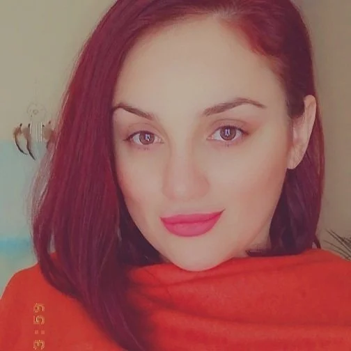 Woman with red hair and red lipstick wearing an orange top, smiling at the camera.