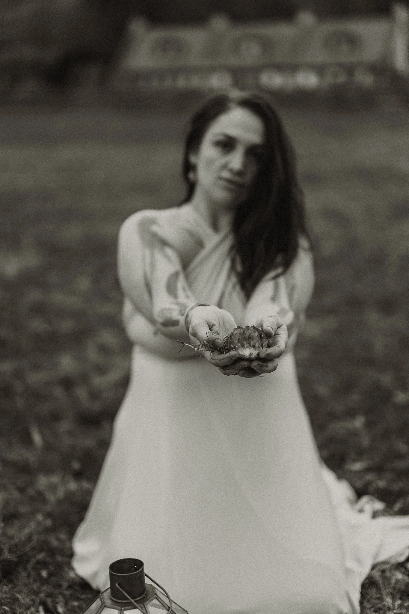 Woman in a white dress kneeling outdoors, holding a small object with soil in her hands, blurred background.