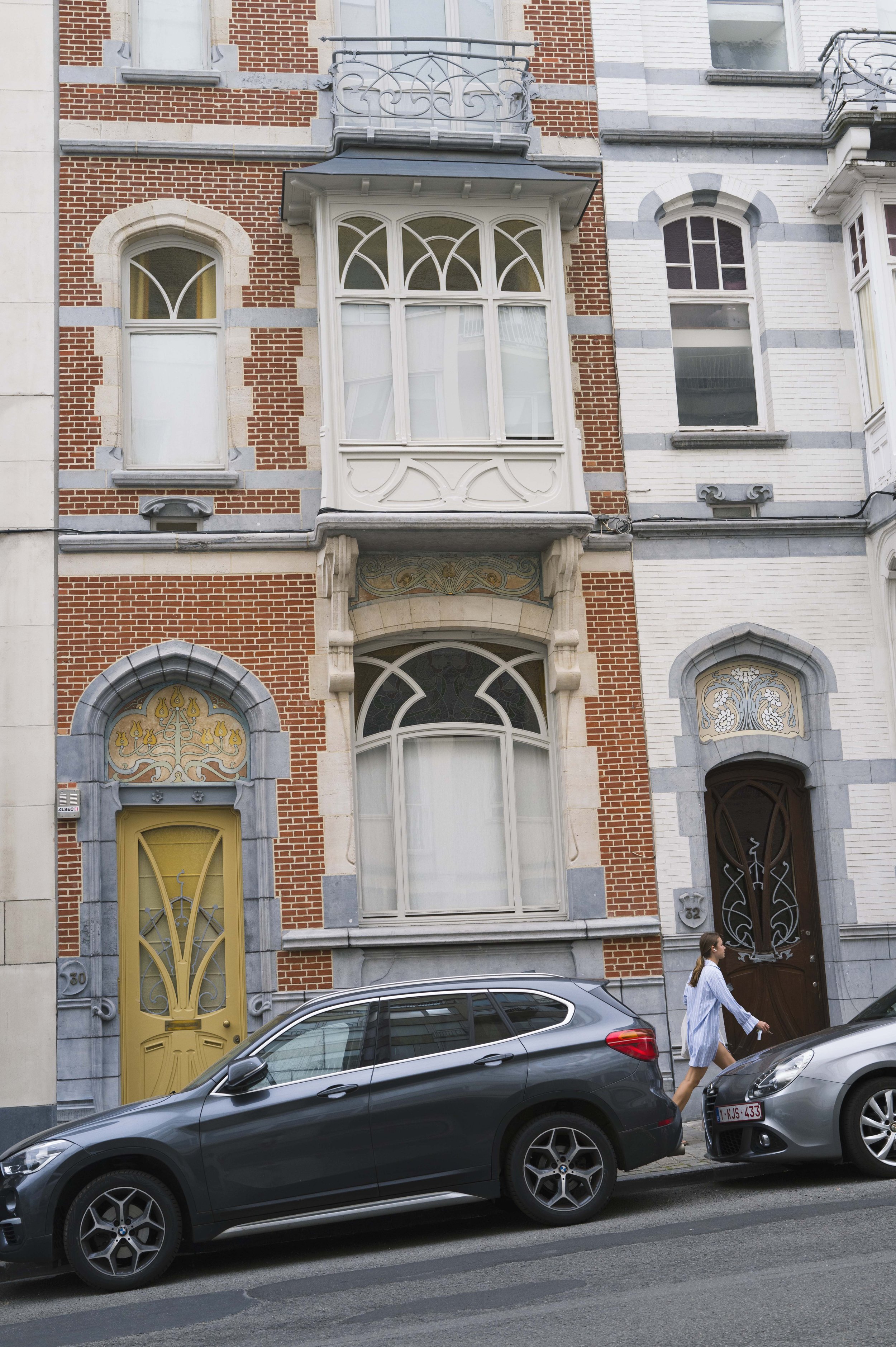 Rue urbaine avec des bâtiments de style européen en pierre et briques, fenêtre avec ornementation en fer forgé, porte d'entrée colorée, deux voitures stationnées sur la route, femme marchant sur le trottoir, chaussée mouillée.
