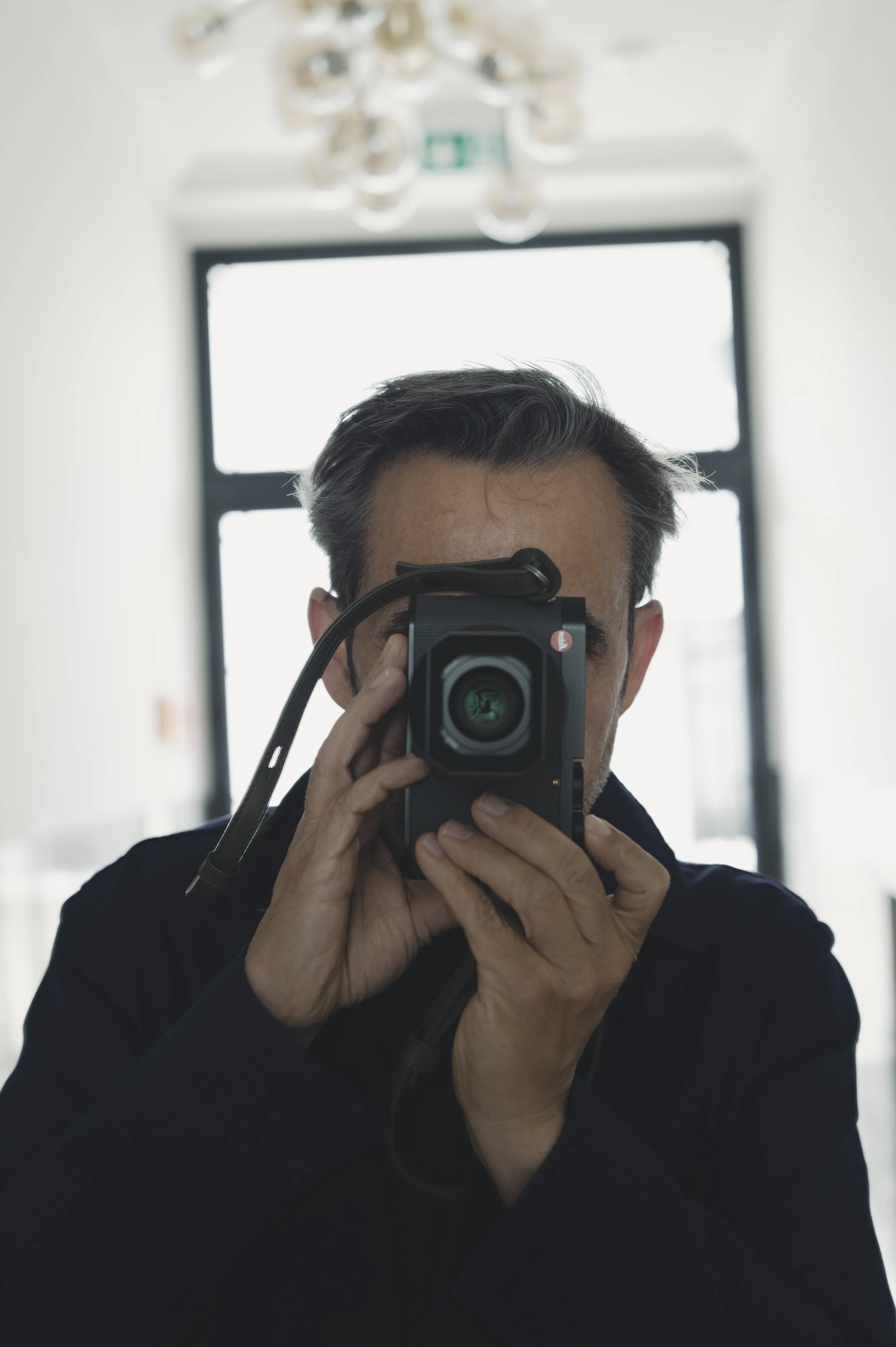 Un homme se prenant en selfie avec un appareil photo noir dans une pièce lumineuse, avec un grand miroir derrière lui et une jonction de plafond en cristal.