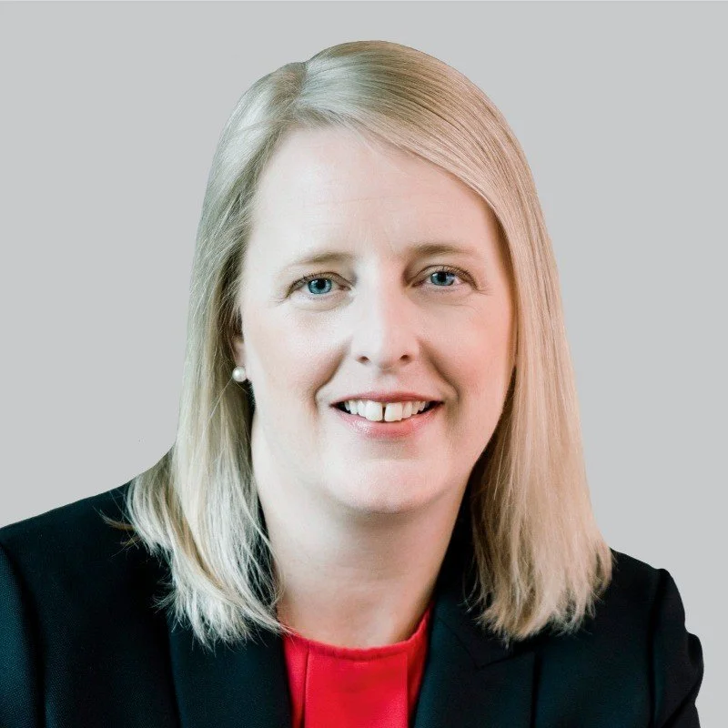 A young woman with curly hair, glasses, and hoop earrings, smiling in a well-lit room with a blurred background.