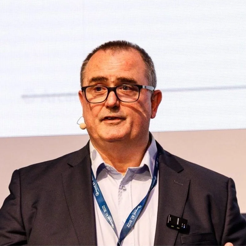 A black-and-white photo of a man in a suit and collared shirt, smiling softly, in an indoor setting with blurred background.