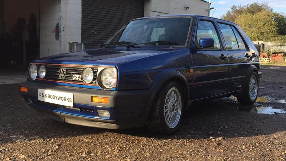 A blue Volkswagen Golf GTI parked outside a garage. The car features a classic design with round headlights and alloy wheels. A sign on the front reads 'C & S Bodyworks.'