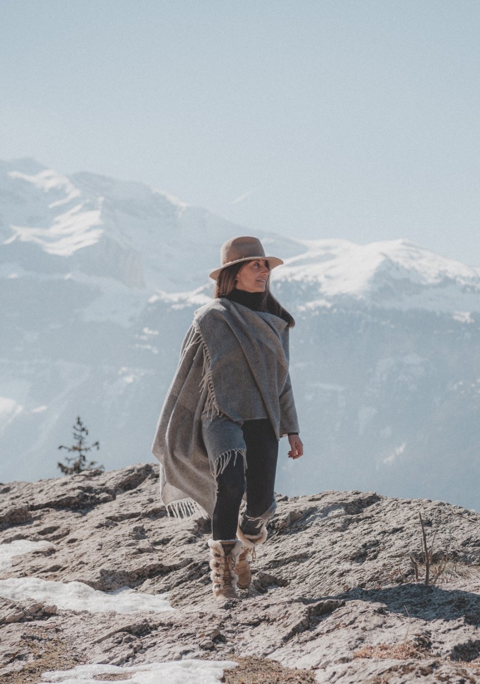 Femme portant un chapeau et une cape en laine, marchant sur une roche en montagne enneigée.