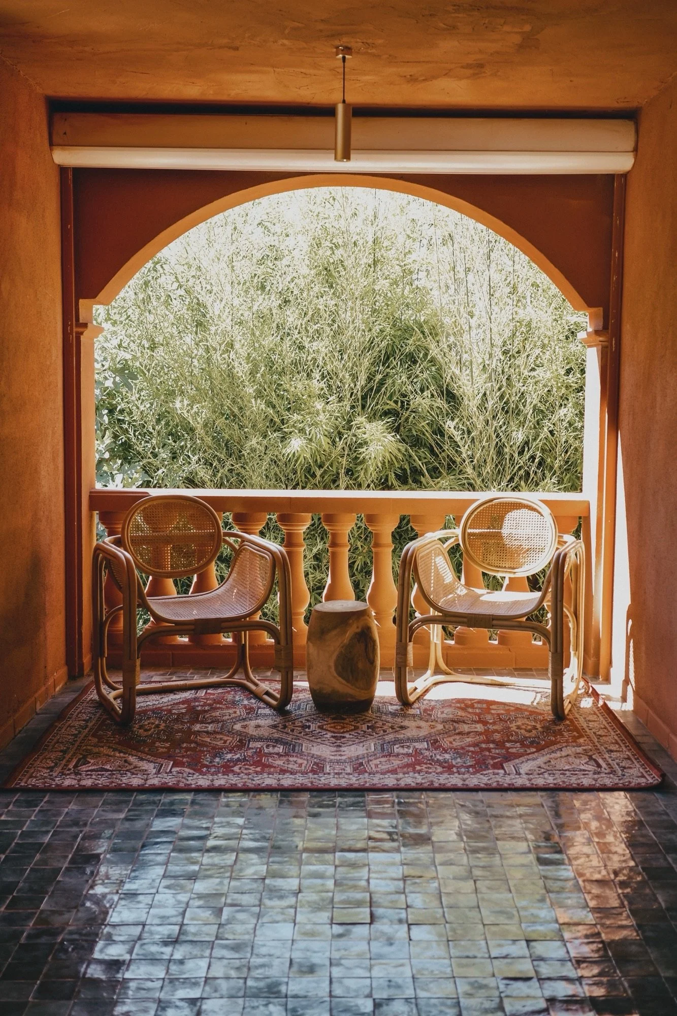 Un balcon avec deux chaises en rotin, un petit tabouret en pierre, un tapis rouge et un grand arbre vert en arrière-plan.