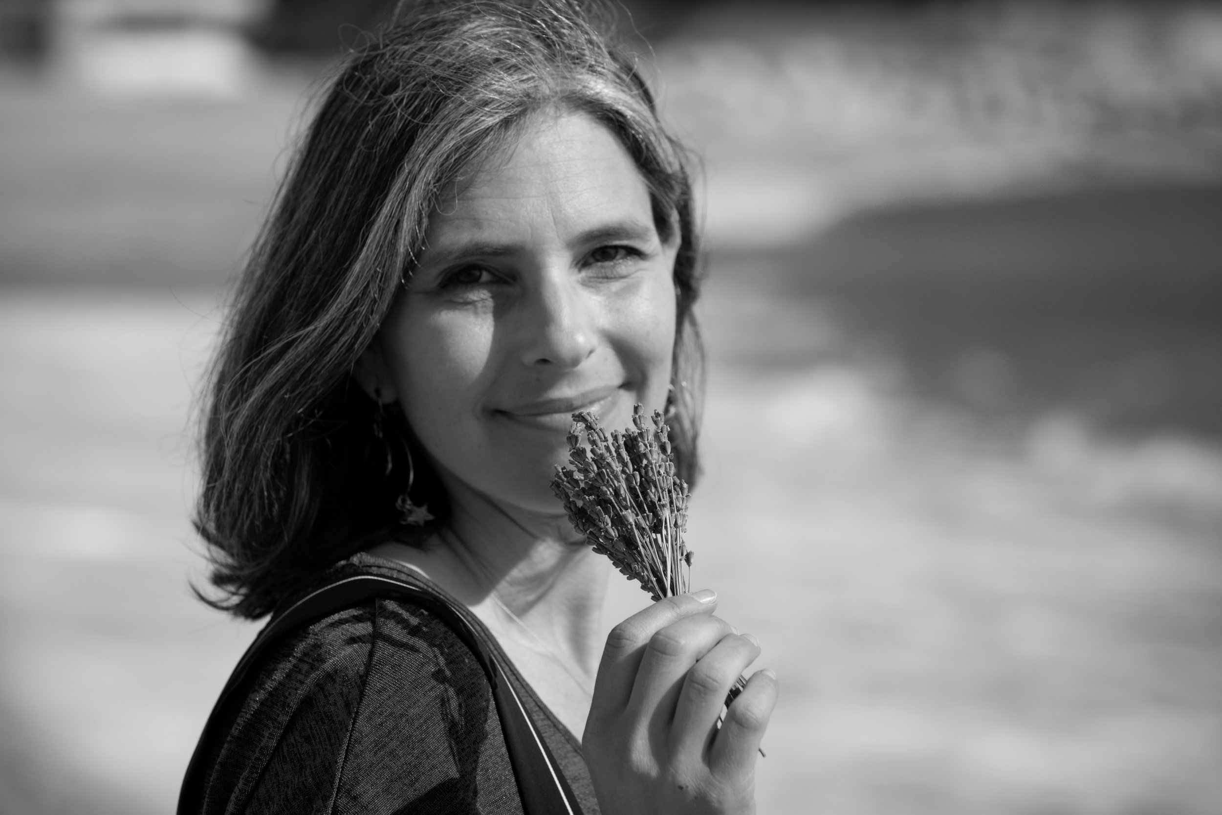Une femme souriante avec des cheveux longs tient un bouquet de lavande près de sa bouche, sur fond flou.