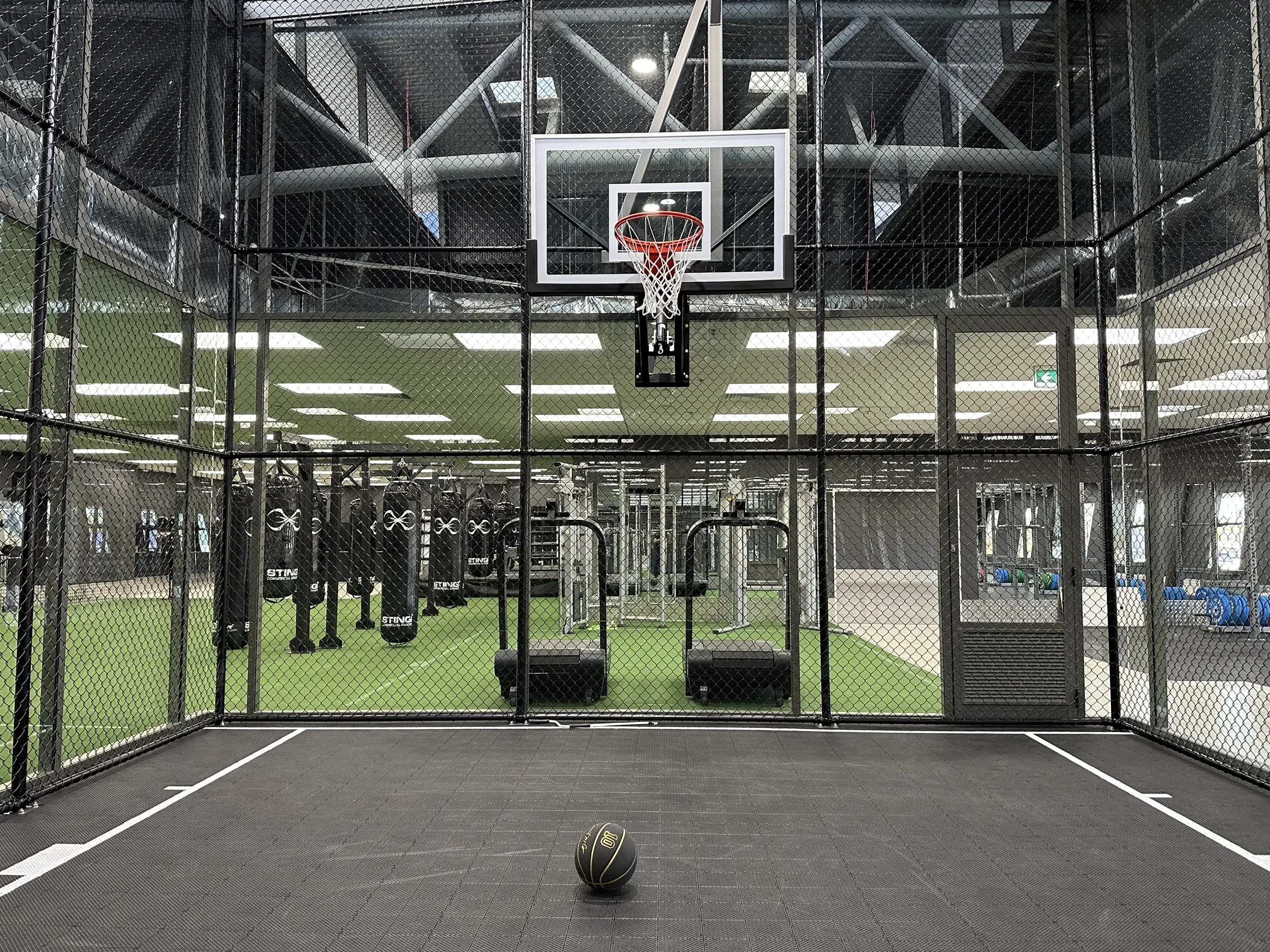 Indoor gym with a small basketball court, a basketball hoop, punching bags, and weight equipment in the background. A black and gold medicine ball is on the court, enclosed by a metal fence.