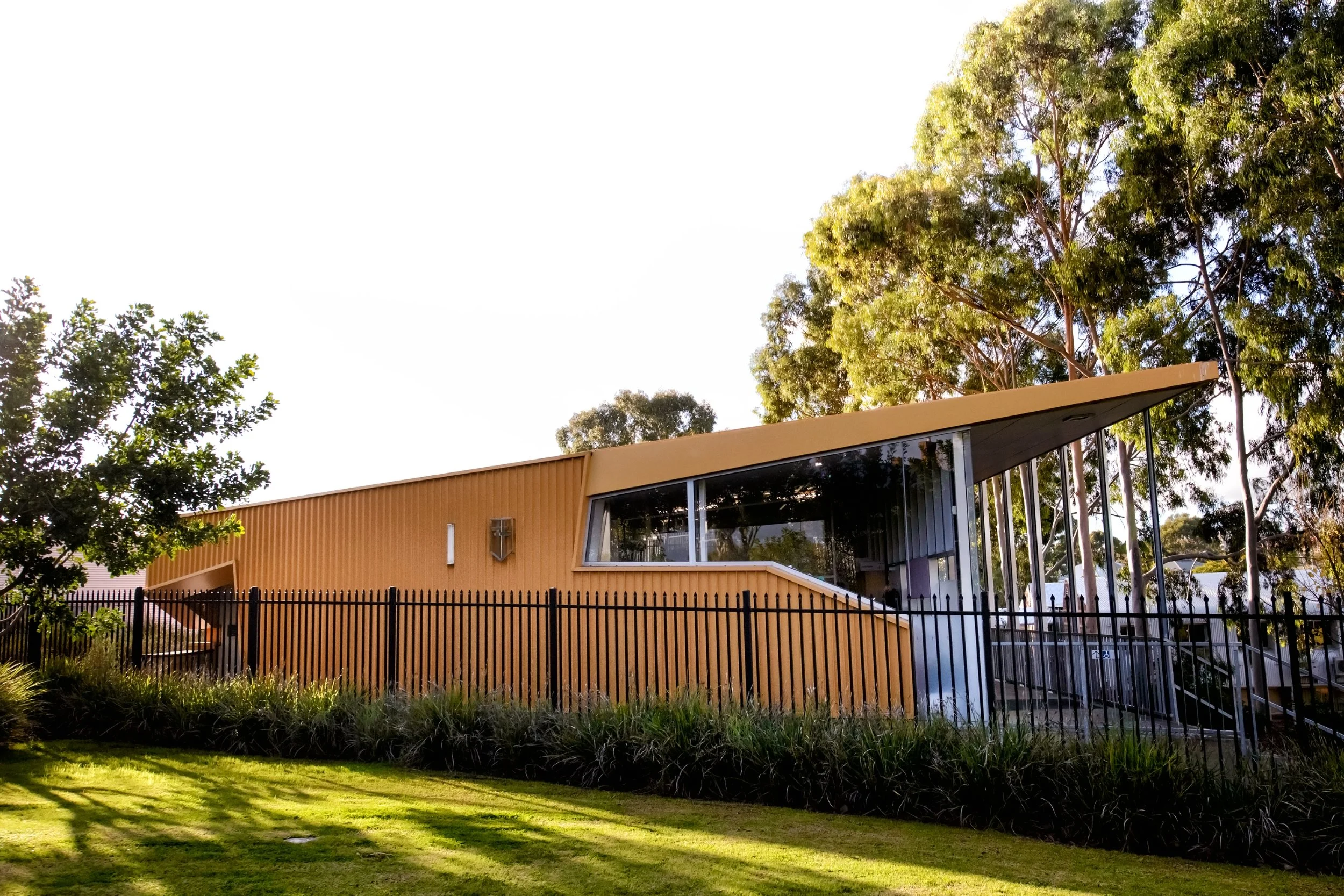 Modern building with a brown exterior, large glass windows, and a sloped roof, surrounded by green trees and grass.