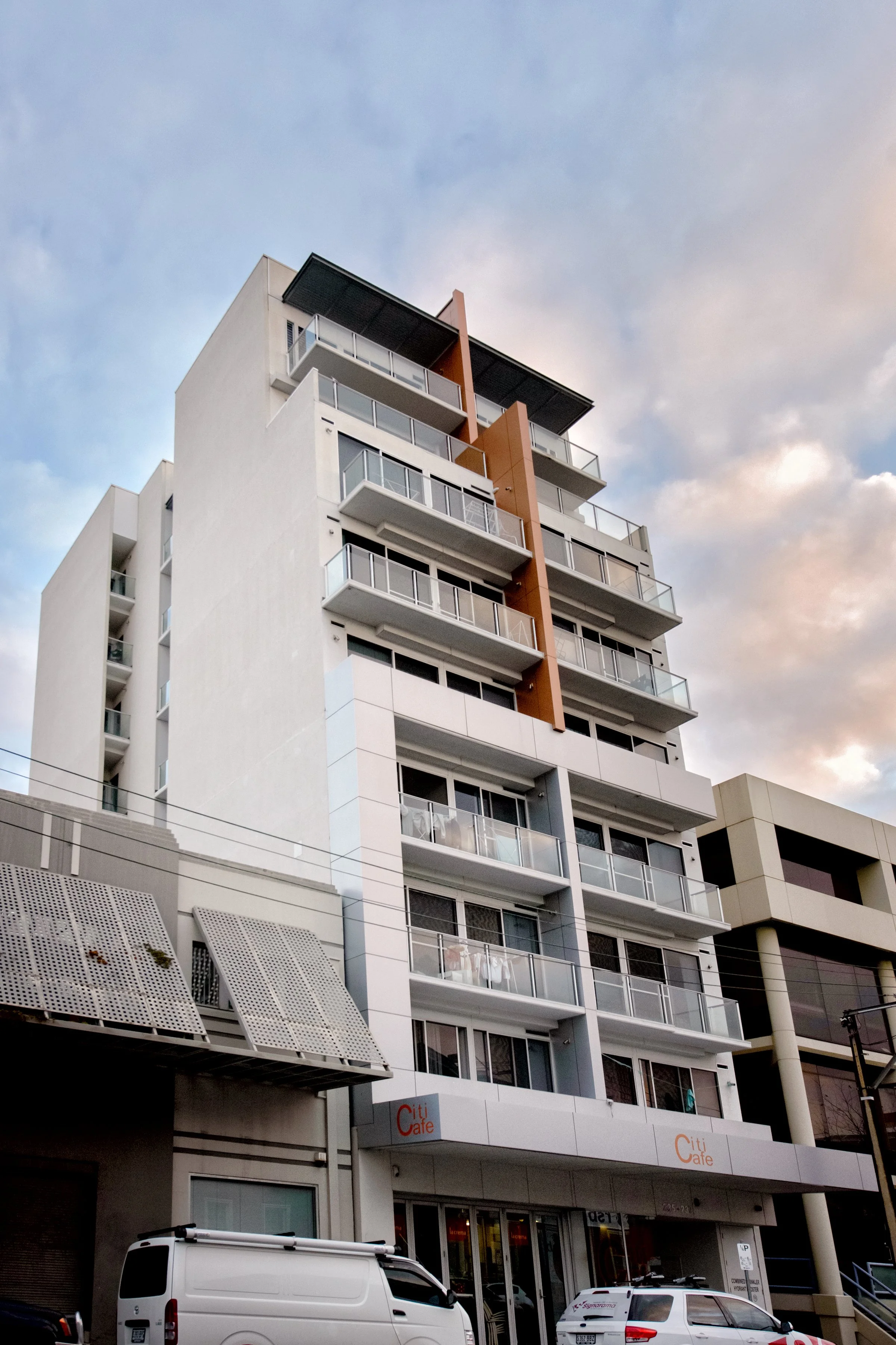 Multi-story modern residential building with balconies and glass railings, adjacent to a street with parked cars and a cafe on the ground floor.