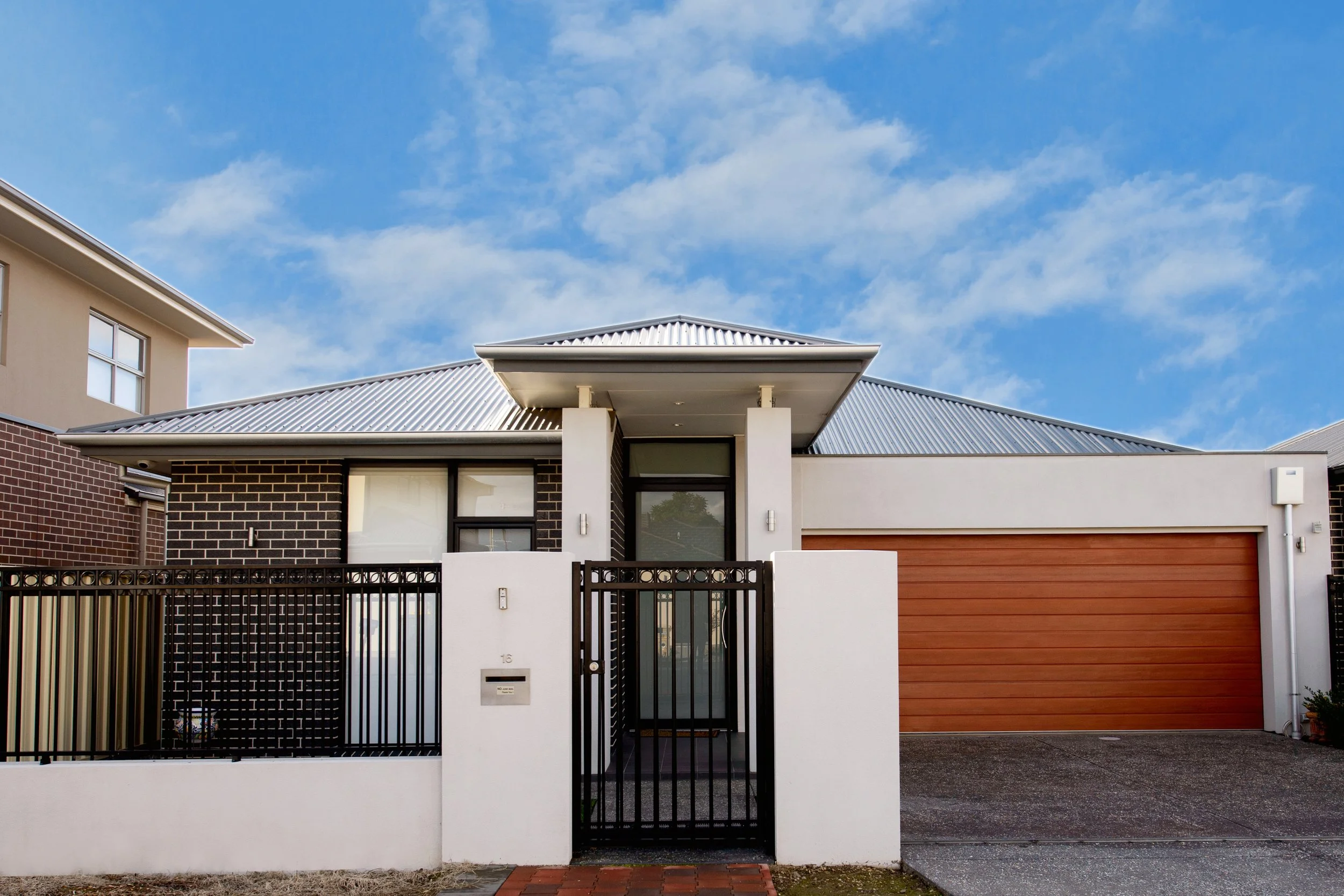 Modern house with a black metal gate, white walls, brown garage door, and a metal roof, under a partly cloudy blue sky.