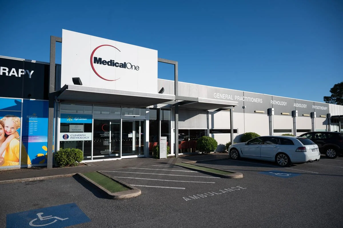 Exterior view of Medical One, a healthcare facility, with parking lot and accessible parking spaces, clear signage for general practitioners and various medical services, and a blue sky in the background.