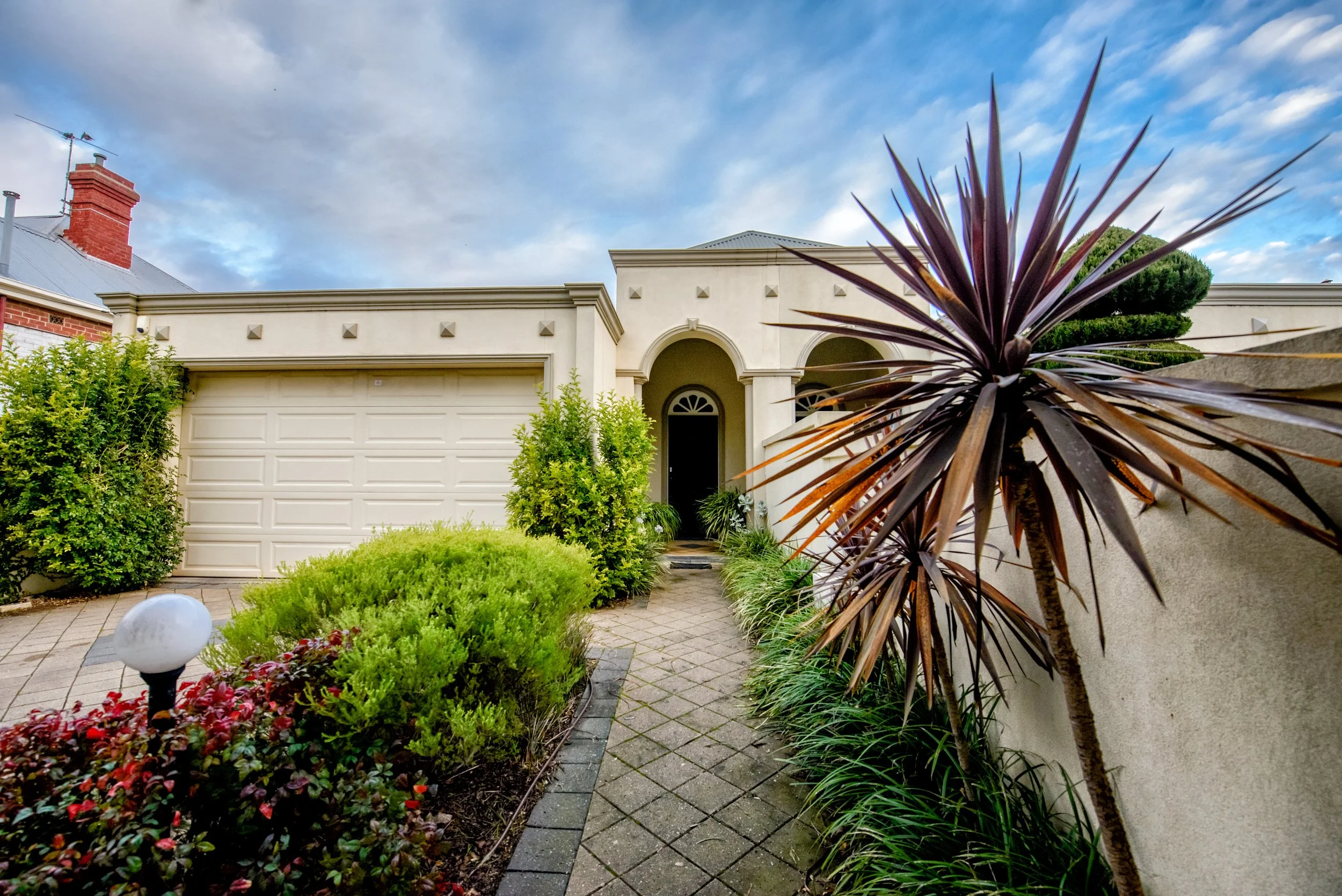 Front entrance of a modern house with a paved walkway, lush green bushes, a large purple-leafed plant, and a closed garage door under a cloudy sky.