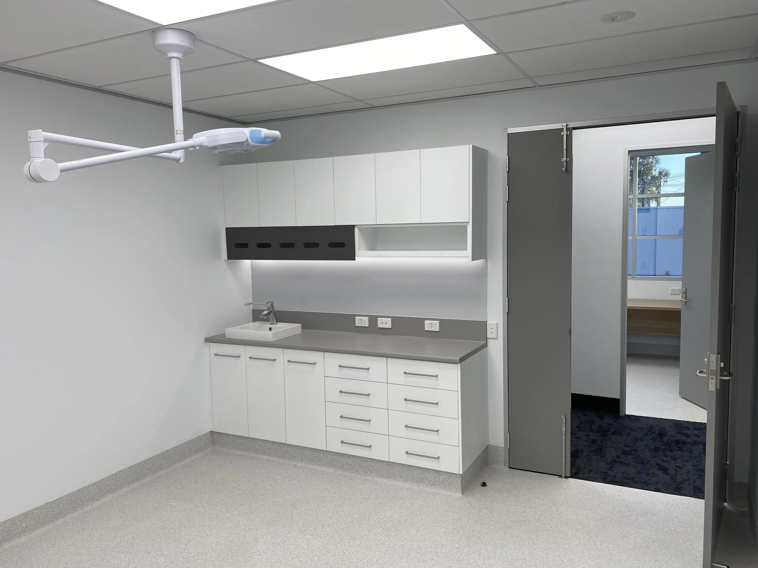 Empty hospital or medical clinic room with a medical light on the ceiling, white cabinets, a small sink, and an open door leading outside.