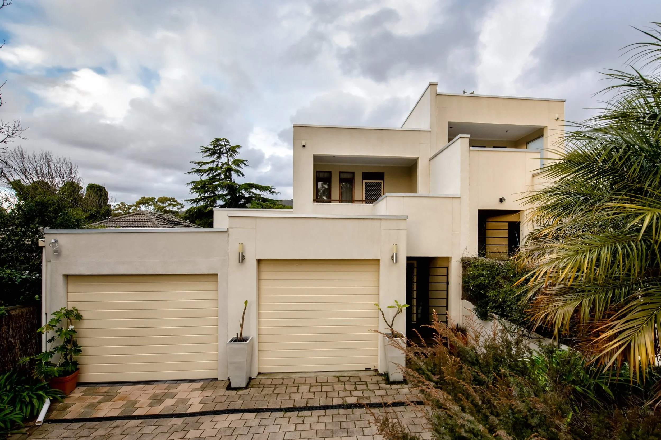 Modern multi-story white house with a garage door, small garden, and overcast sky.