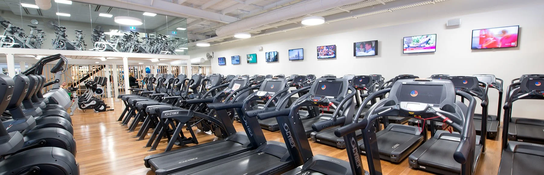 Empty gym with treadmills, ellipticals, and exercise bikes, large monitors on the wall, and a mirrored section with stationary bikes in the background.