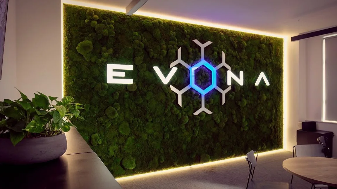Modern office interior with a green wall featuring moss and a glowing logo that reads "EVONA" with a blue-lit hexagon and molecular design